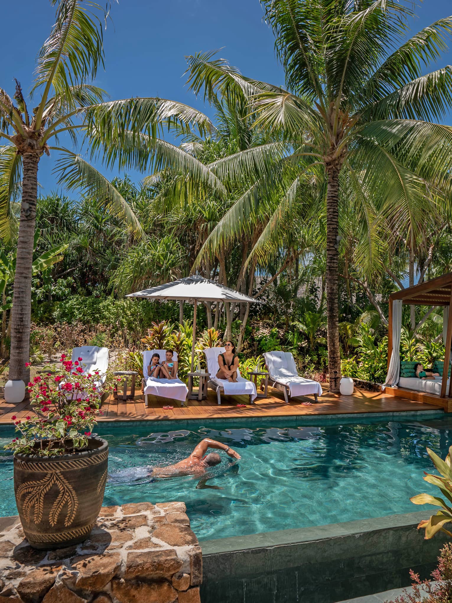A man swims in an outdoor pool surrounded by palm trees, adjacent dek has white lounge chairs and umbrellas
