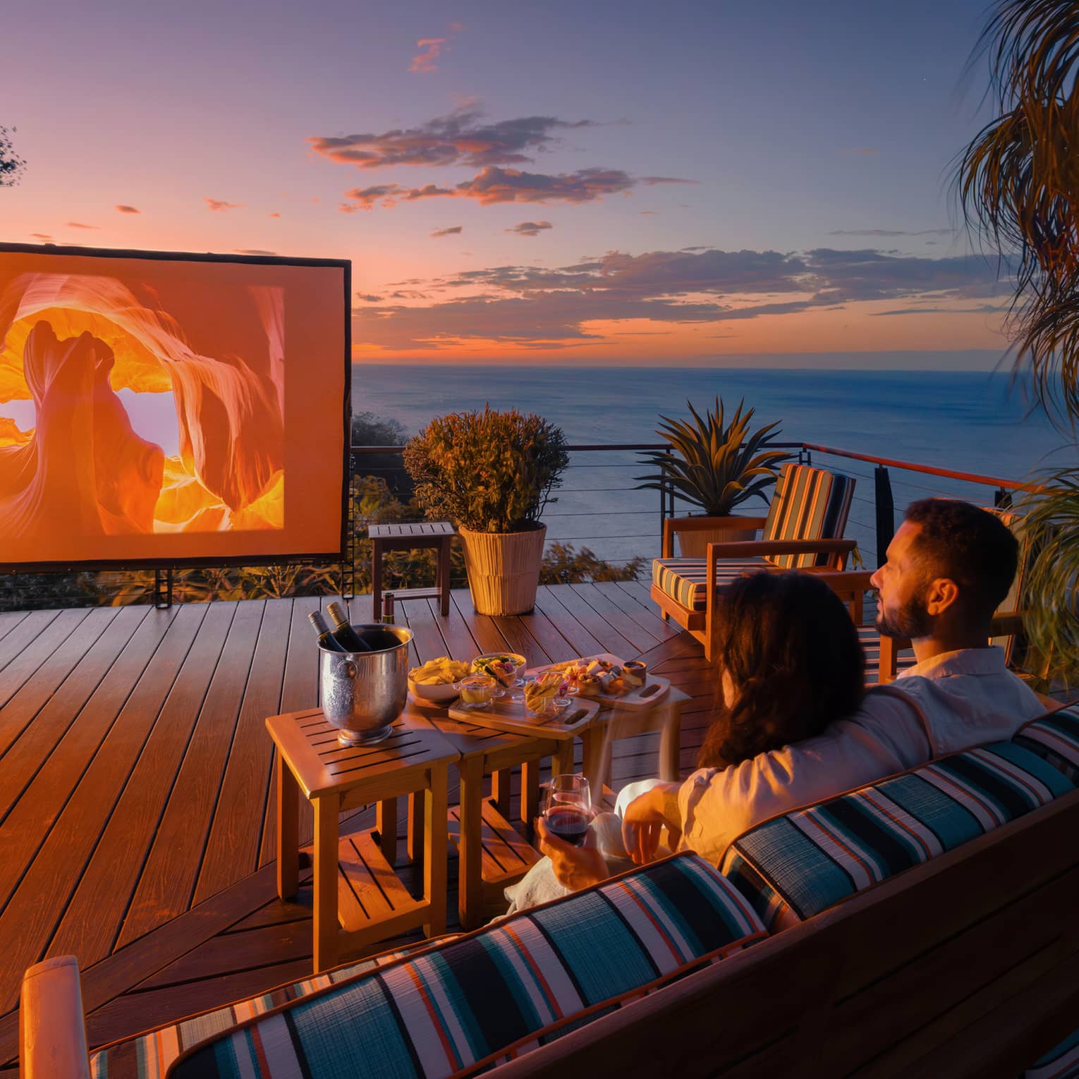 Two people sit on an outdoor sofa with a small table of food infront of them as they watch a movie on a large outdoor screen set up on a private terrace