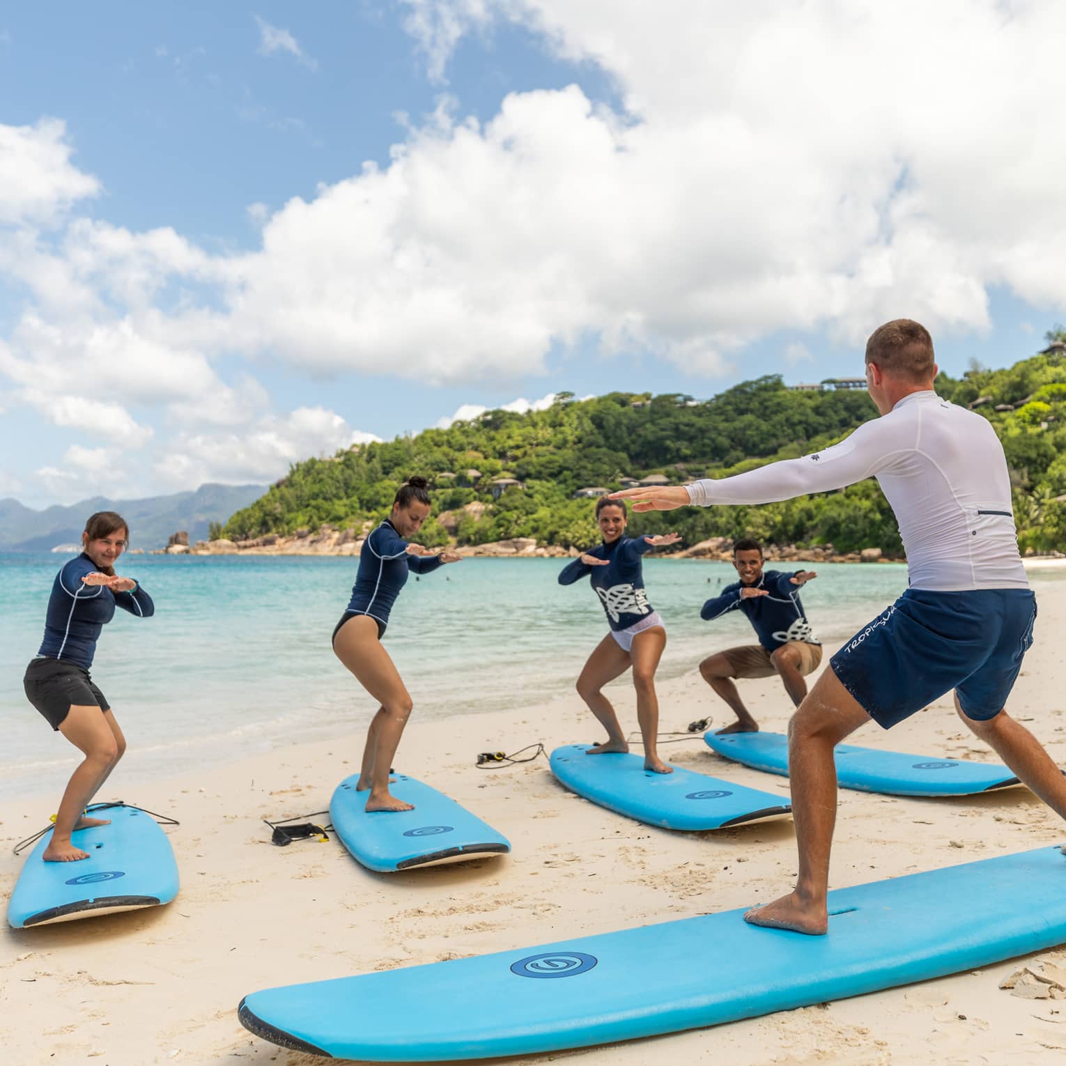 Surfing instructor balances on surfboard on white sand beach in front of row of surfers