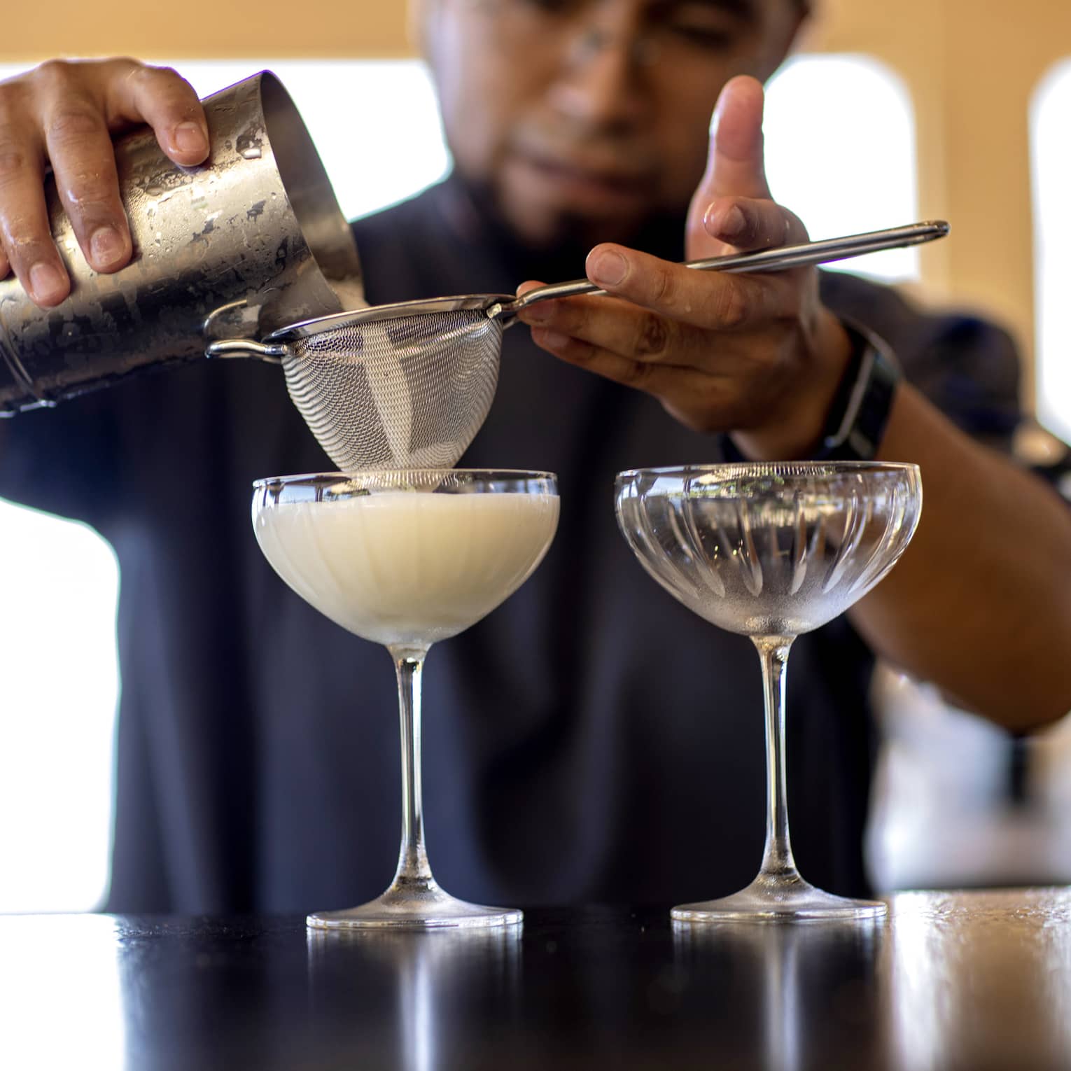 From a metal cocktail shaker, a bartender filters a milky cocktail through a sieve into an etched groove coupe glass. 