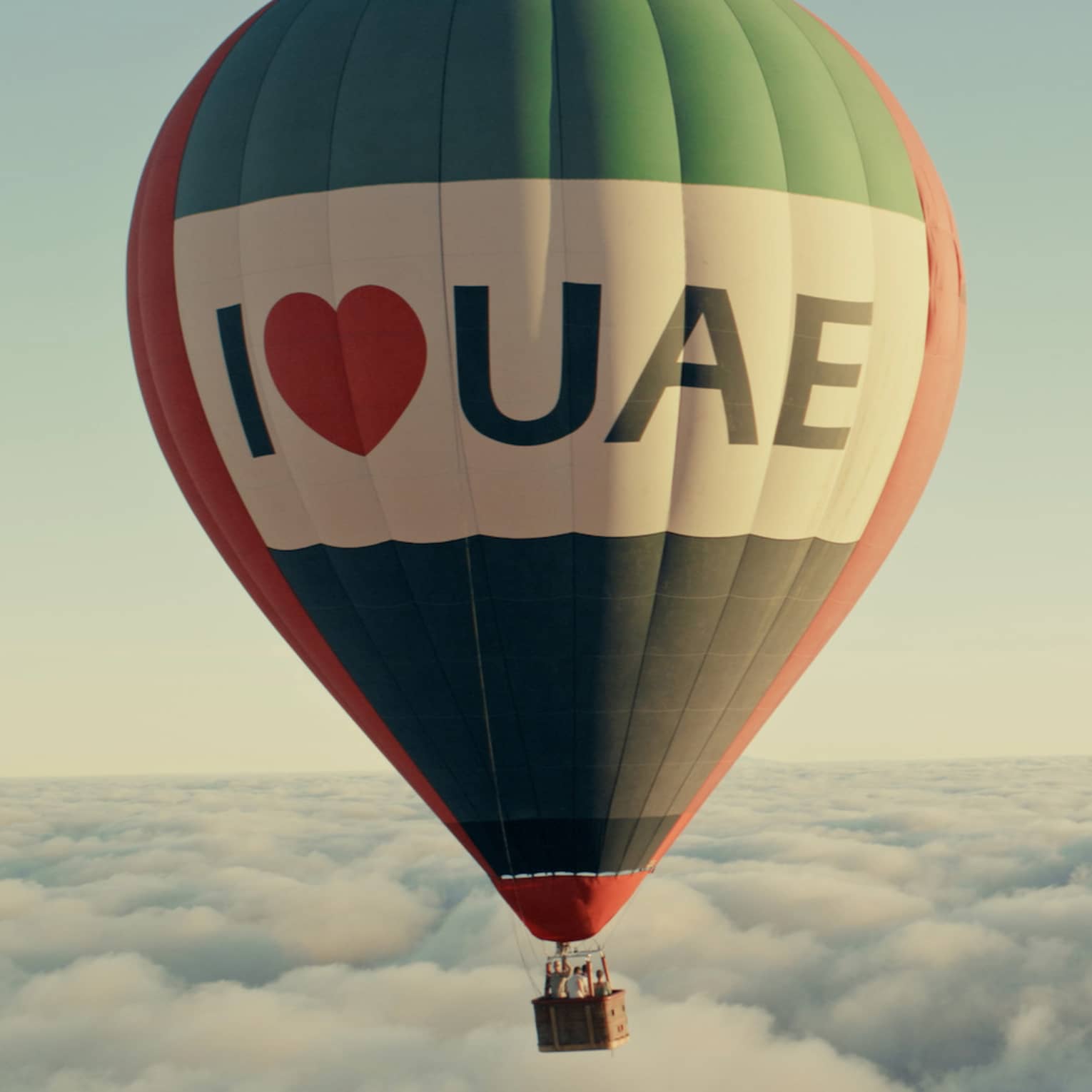 A hot air balloon flies above the clouds, in front of a light blue sky.