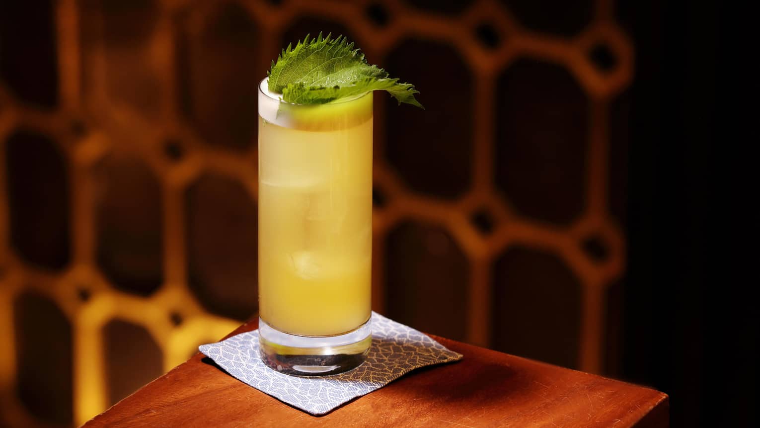A yellow cocktail garnished with a green leaf sits on the corner of a wooden table