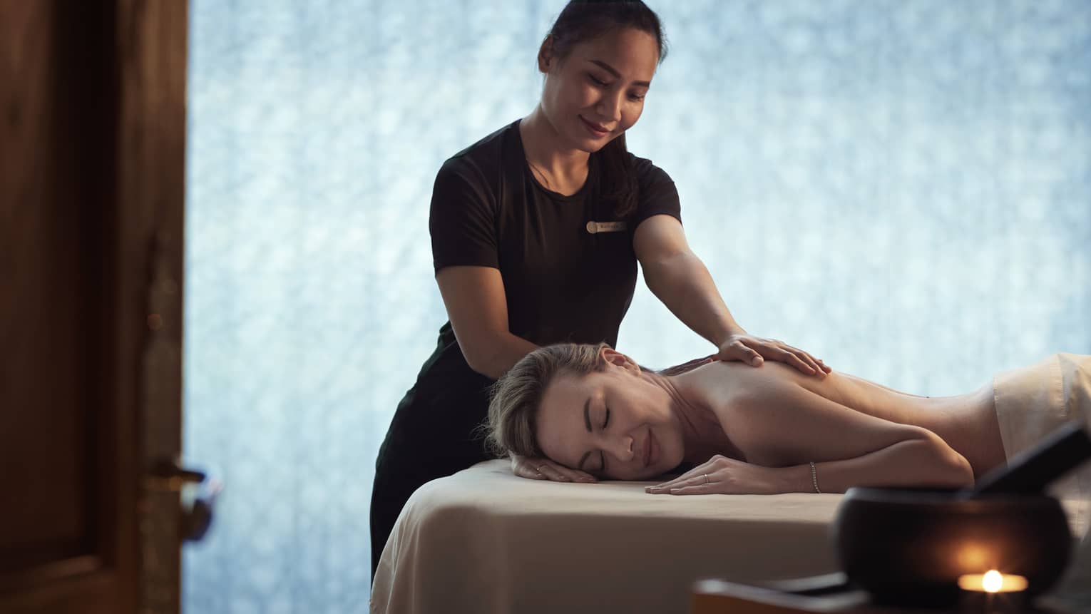 Spa staff massages woman's shoulders as she lays on bed in dark room