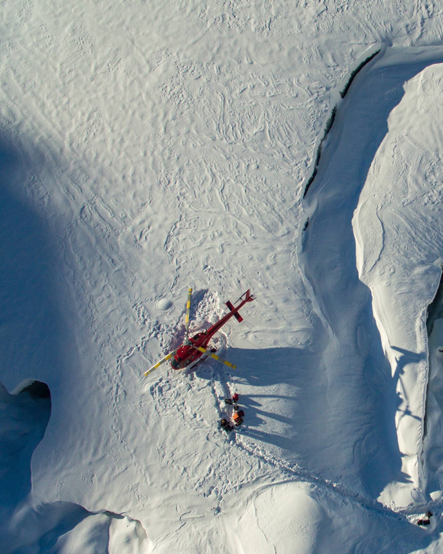 Aerial view of a red helicopter that's landed on a snow-covered mountain with three people walking near it