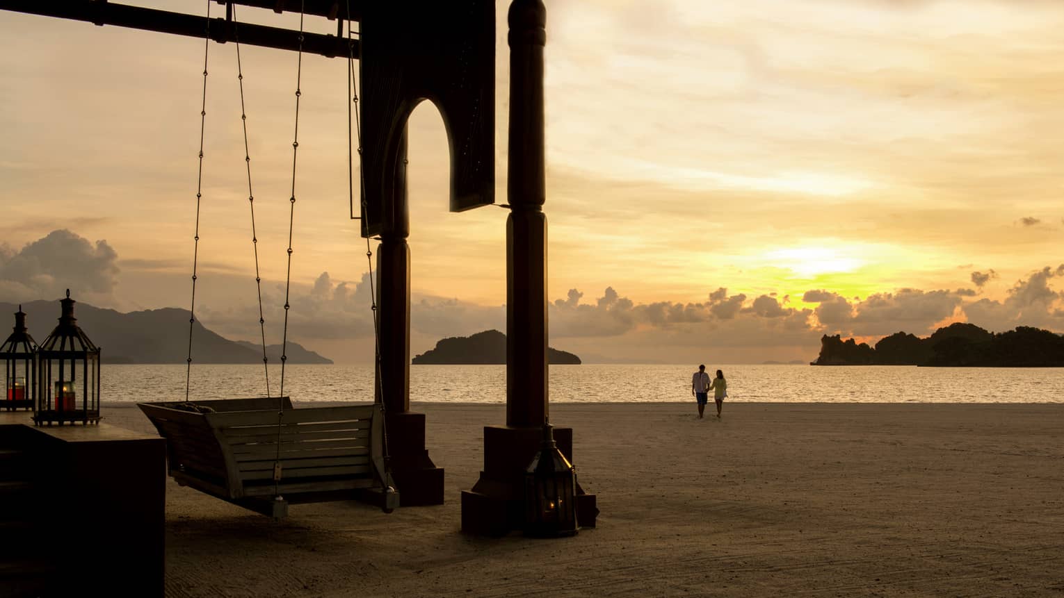 Silhouettes of couple walking along beach past wood swing, pavillion at sunset