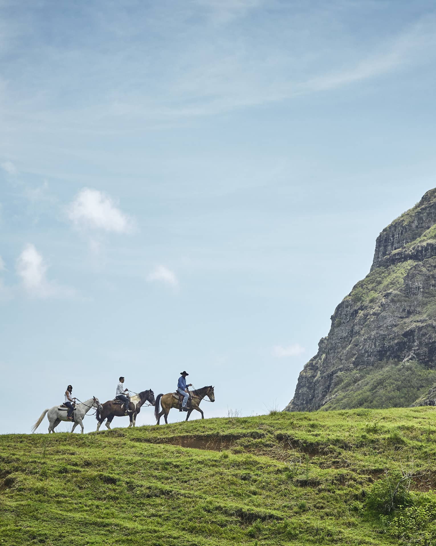 Long side view of three horseback riders on a grassy hill heading toward an imposing verdant cliff under clear blue skies.