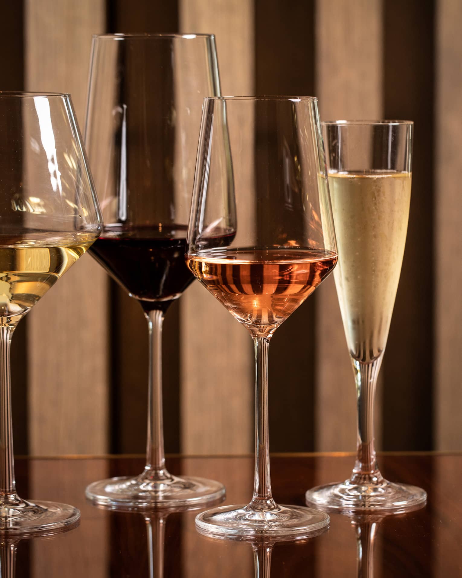Four modern wine glasses holding white, red and rose wine are lined up on a wooden table 