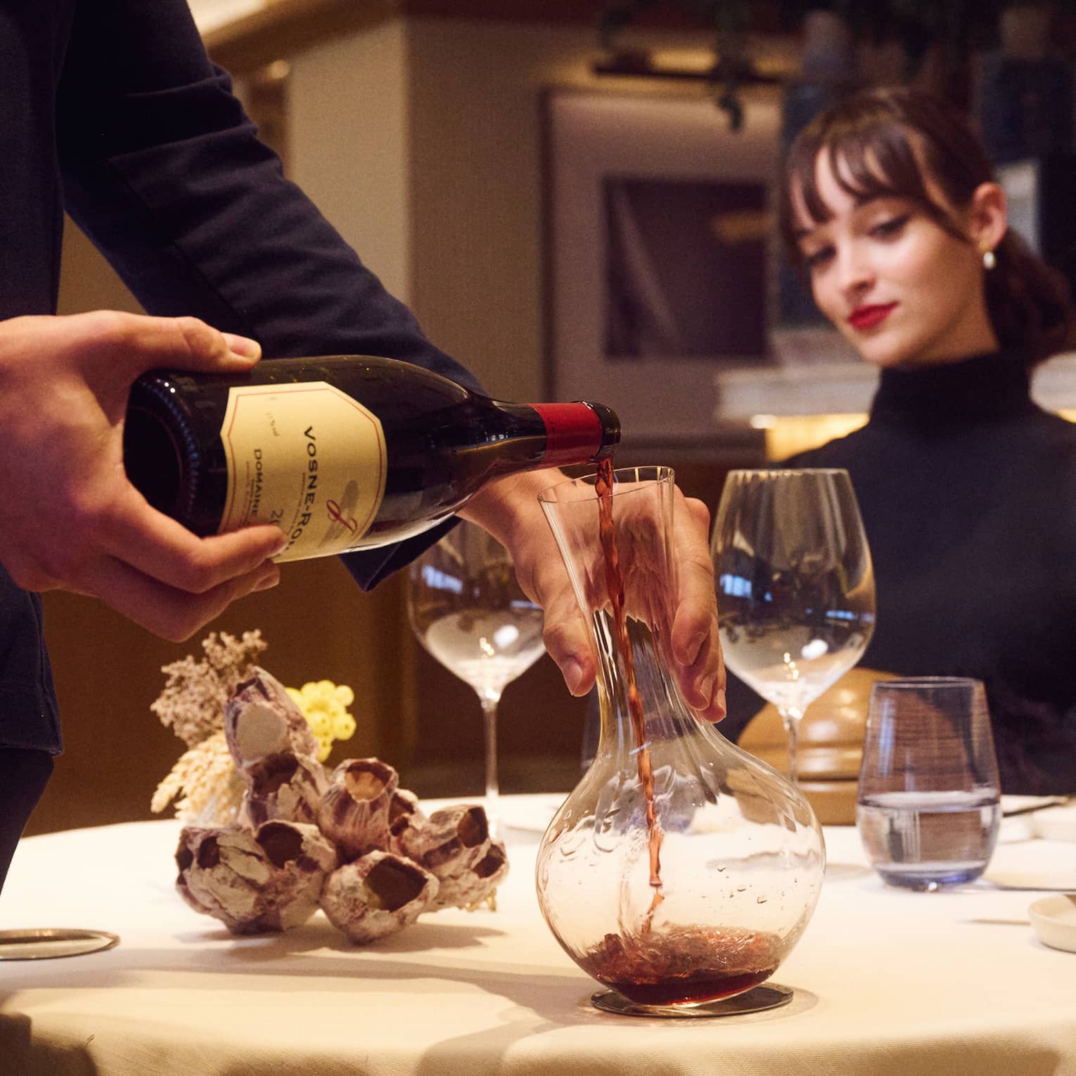 Suited server decants red wine at table with seated guest watching