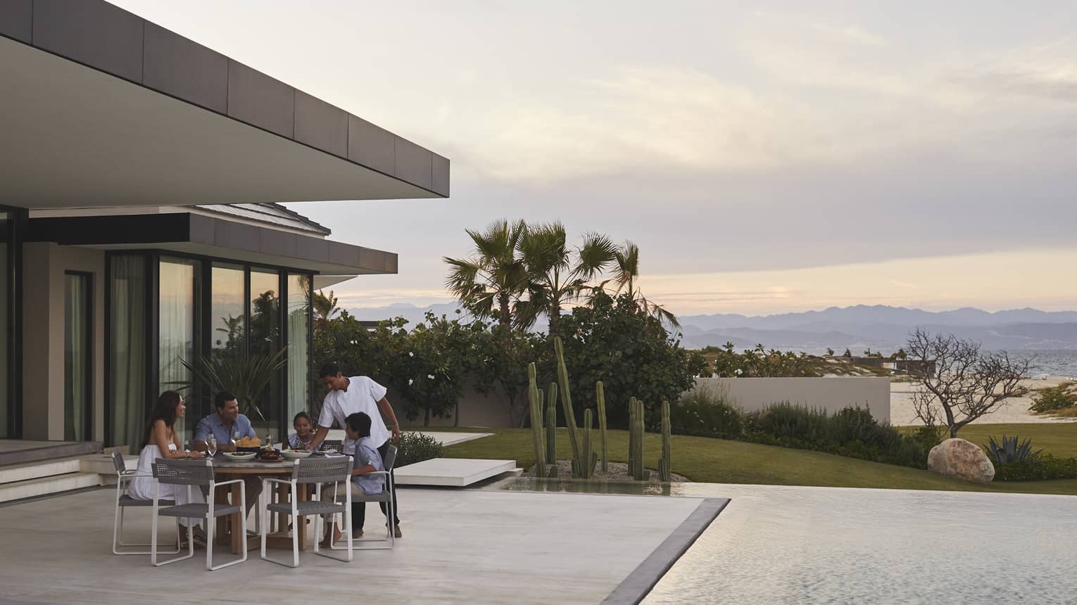 Family dining outdoors on a modern patio by the pool, with a scenic view of mountains and desert plants at sunset, creating a serene and intimate setting.