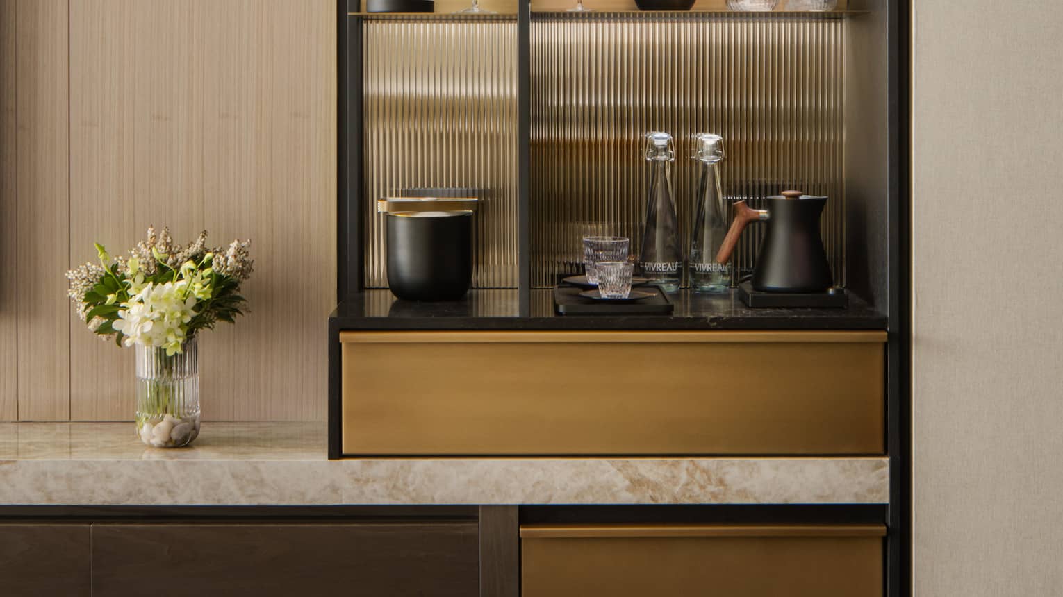 Stylish minibar with glass shelves displaying wine, glasses, and decorative items, next to a TV and vase with fresh flowers