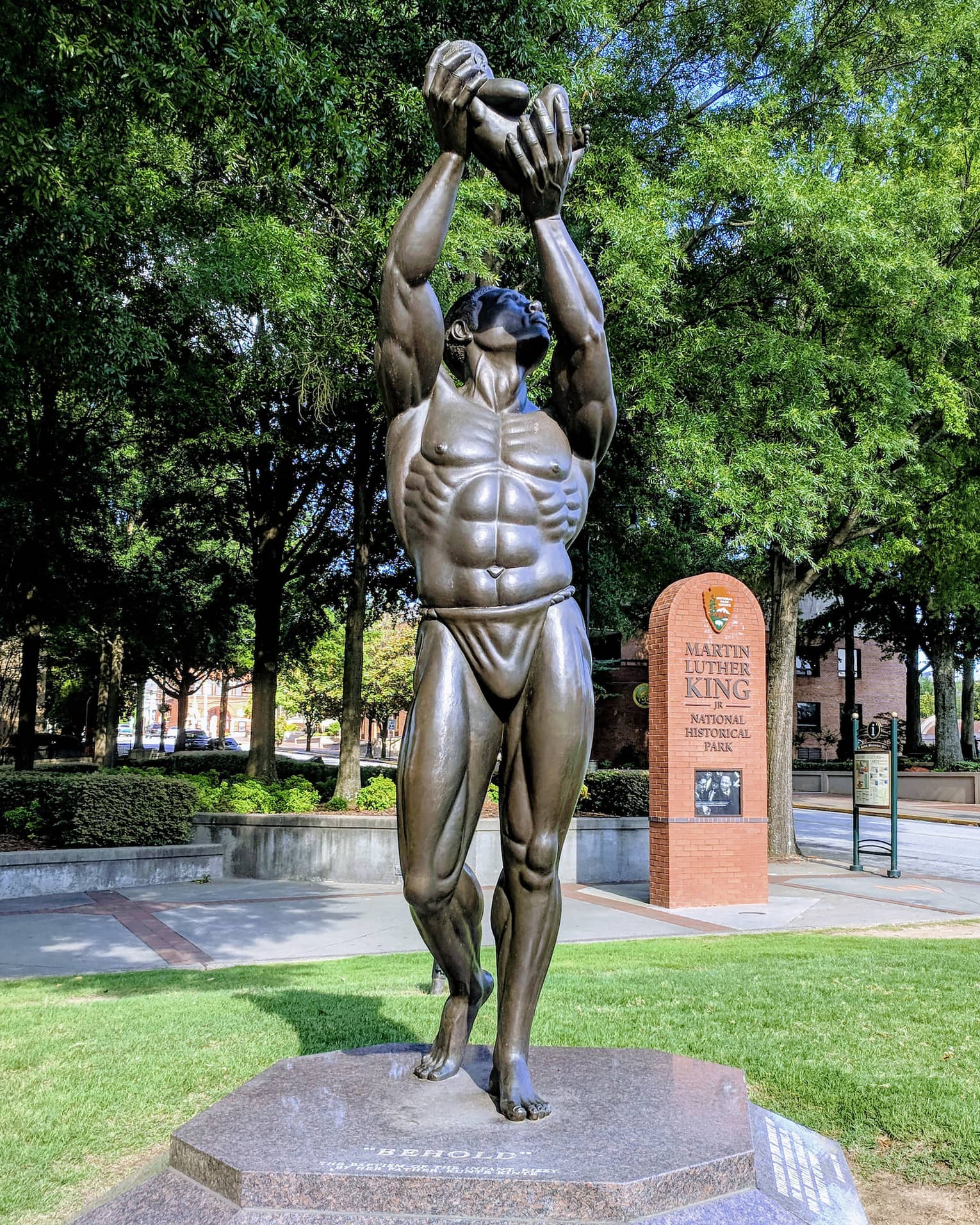 A statue of a man holding something in the air placed in a park with green trees surrounding it.