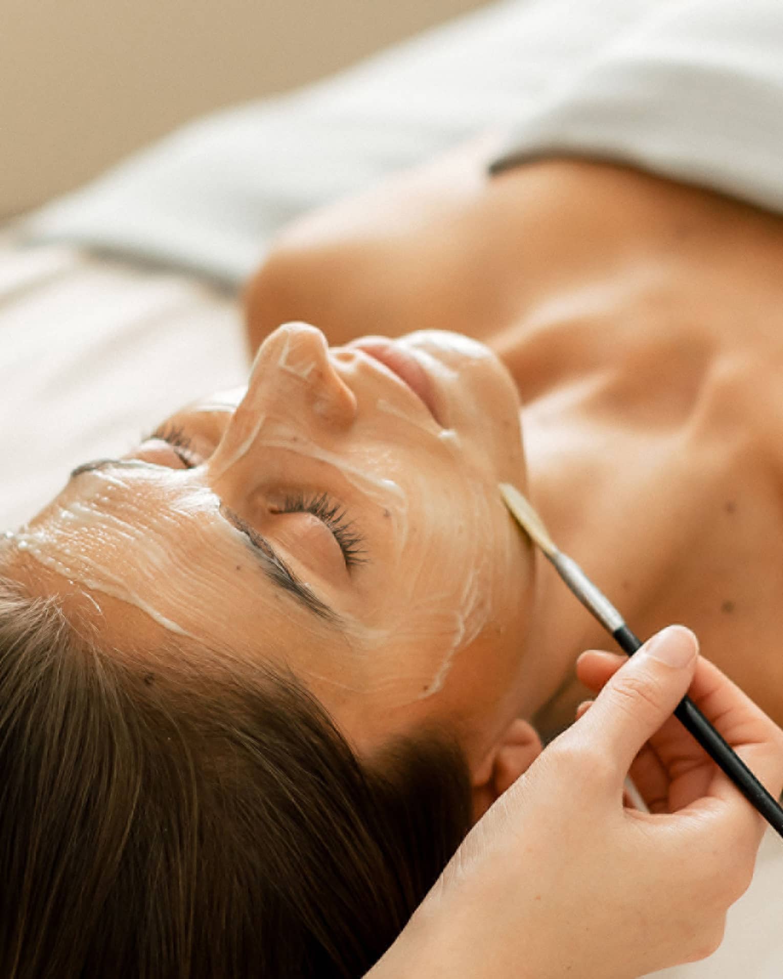 A woman lying on a spa table with eyes closed while a face mask is applied with a small brush