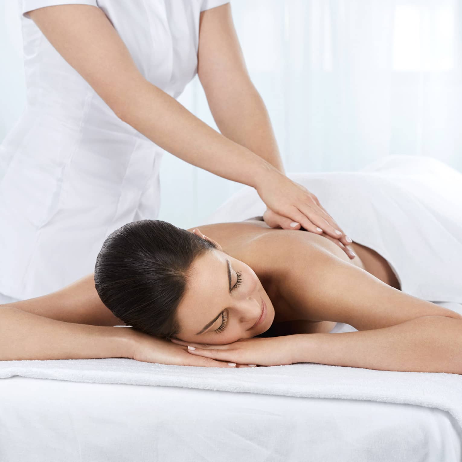 Woman lies under white sheet on massage table as hands massage her bare back