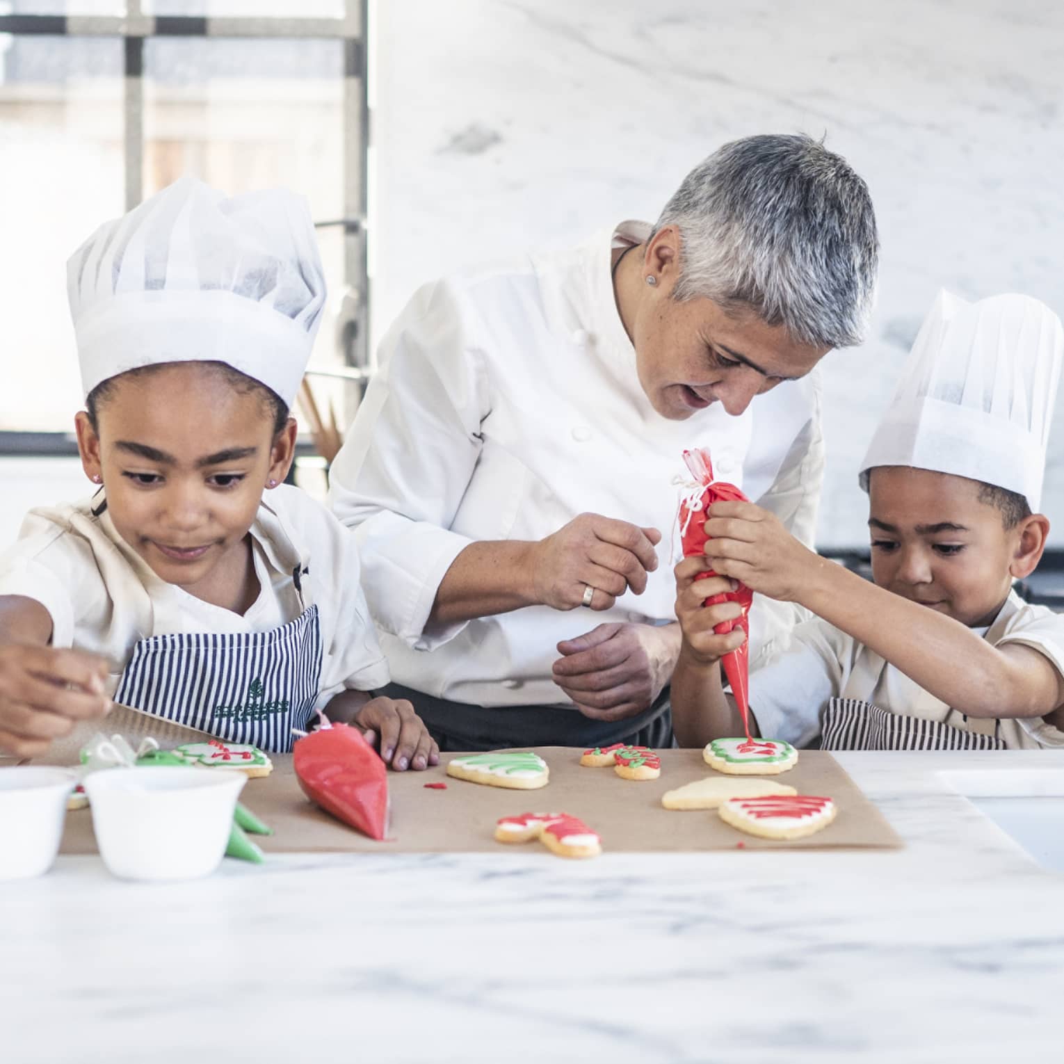 Two children in chef’s hats and aprons in a stark, white kitchen; a chef watches closely as one pipes icing onto a cookie.
