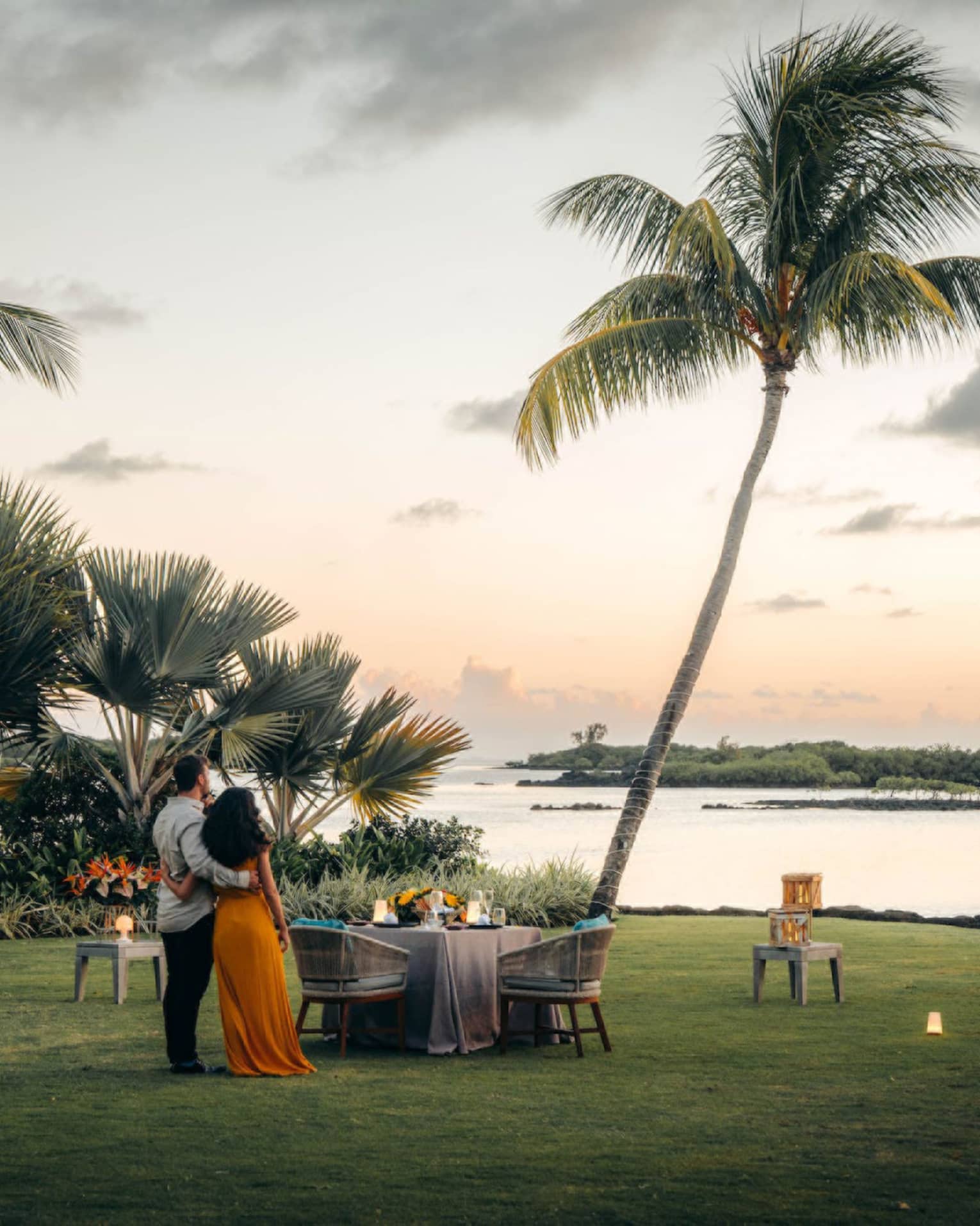 A couple stand on the shore, arms around each other, gazing toward the ocean; set table with two chairs beside swaying palms.