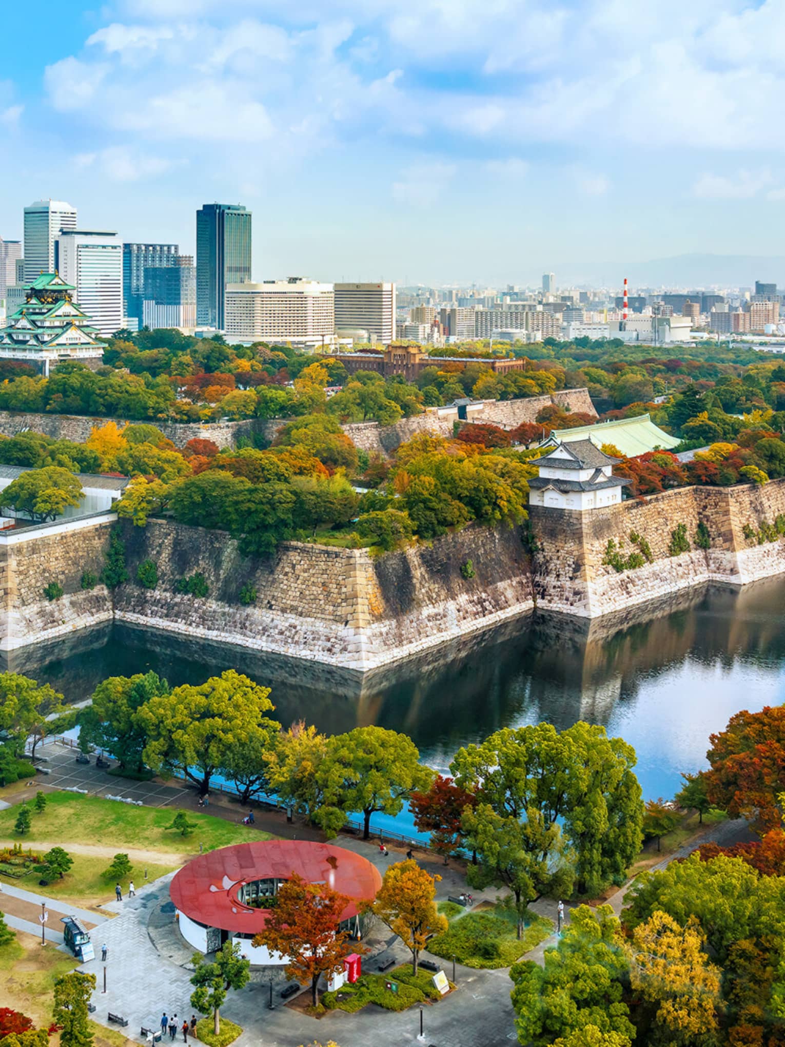 Osaka Castle rises amid lush autumn-coloured trees within a stone fortress surrounded by a moat and modern high-rises.