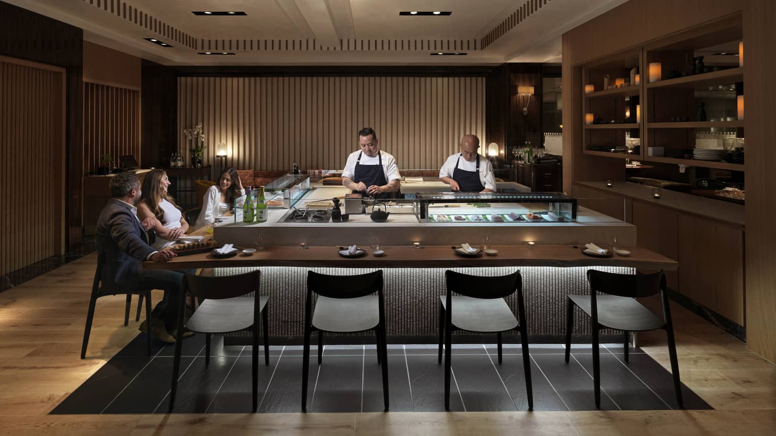 Three guests sit at a sushi counter, engrossed in conversation, while two chefs behind the counter carefully prepare food.