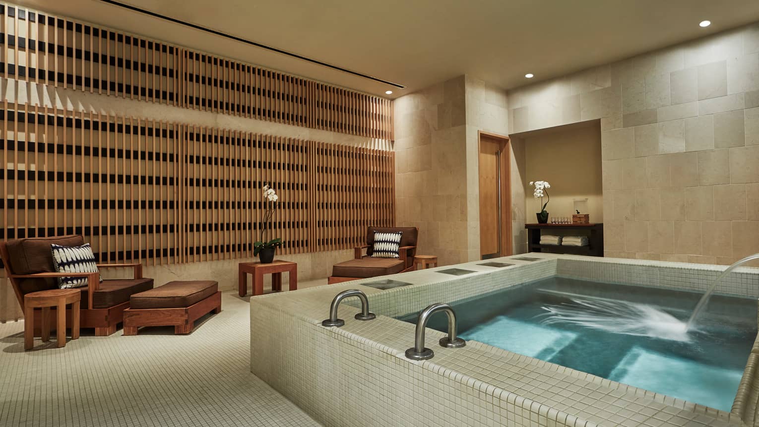 Large square spa tub with water jets in tile room with two brown lounge chairs 