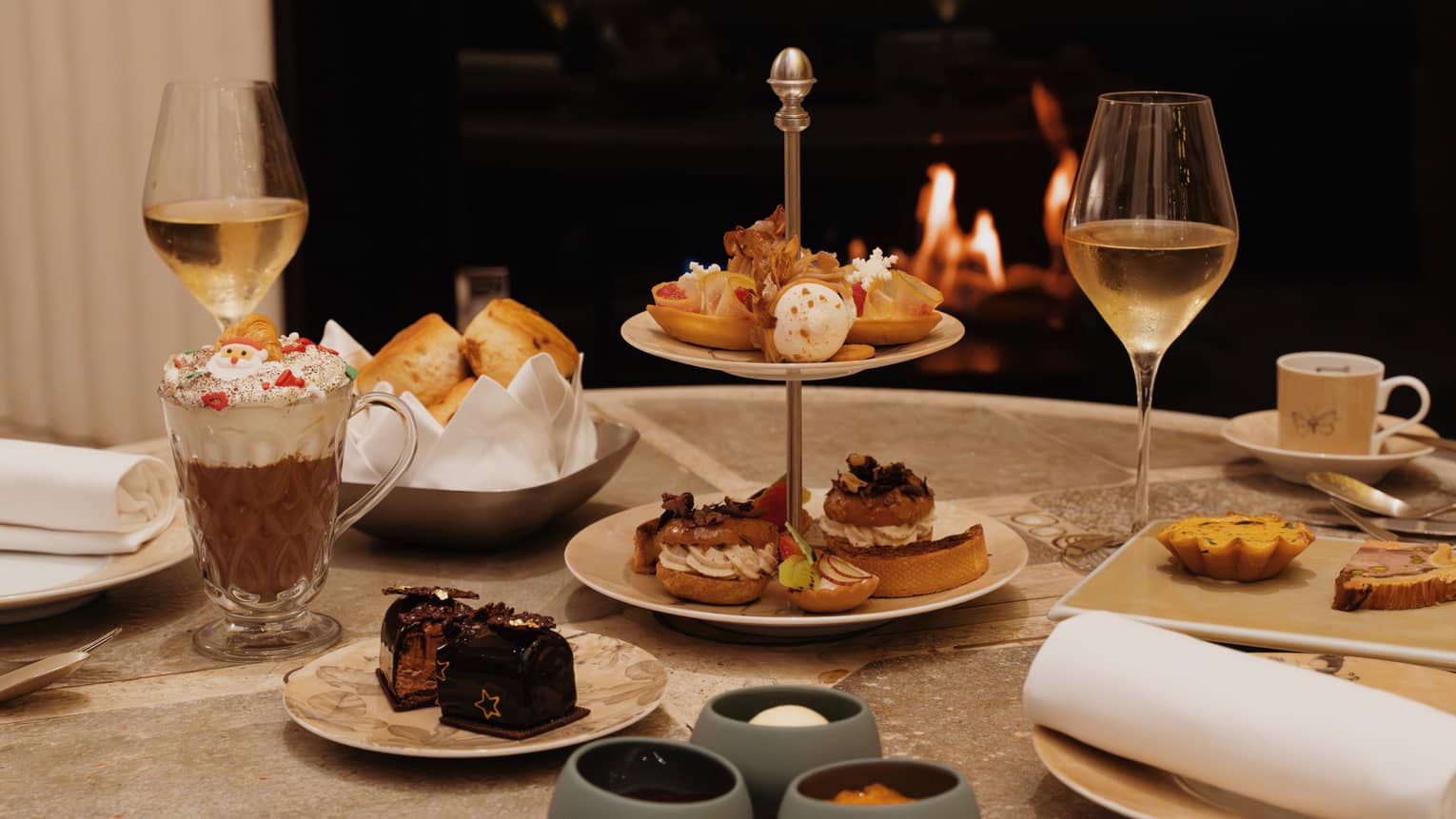 A round table is set for an afternoon tea, with small bites on serving trays, plates and stands, along with beverages. A cozy fire in a fireplace is in the background.