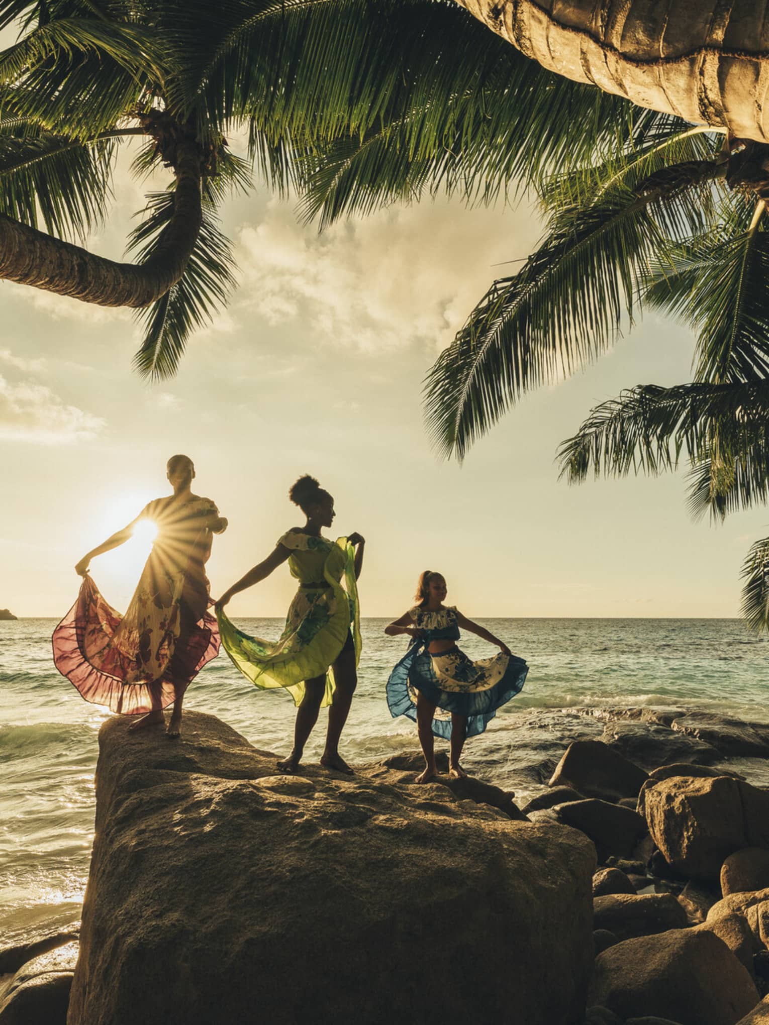 The sun sets over three dancers wearing long, colourful dresses standing on top of rocks by the water
