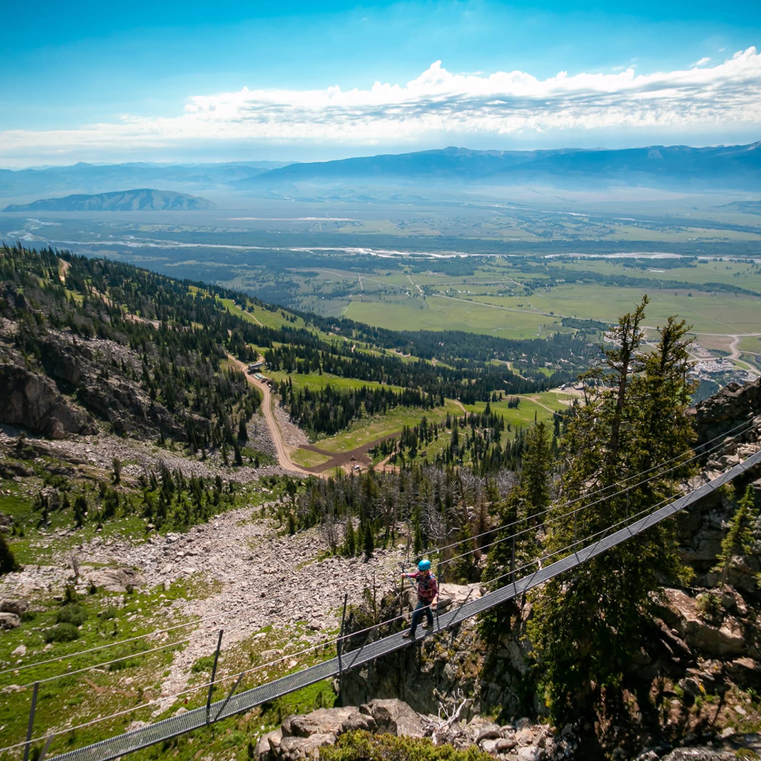 A man walks across a tightrope, a view of the lush green landscape below him 