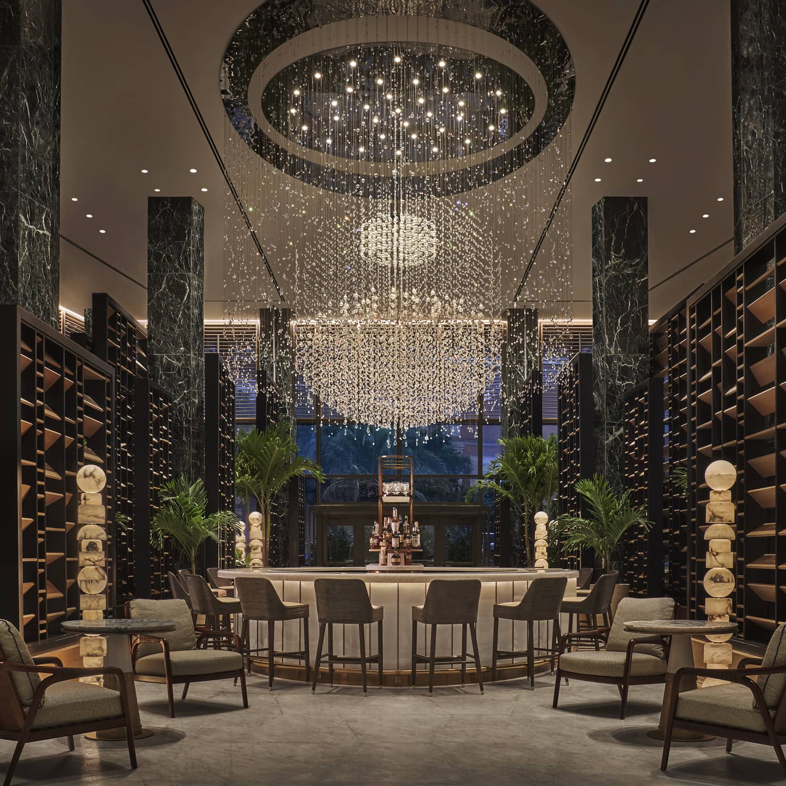 Chandelier Bar with six jade pillars flanked by wooden shelves and potted palms, glass-topped bar in the middle with six cocktail chairs, giant chandelier hanging overhead