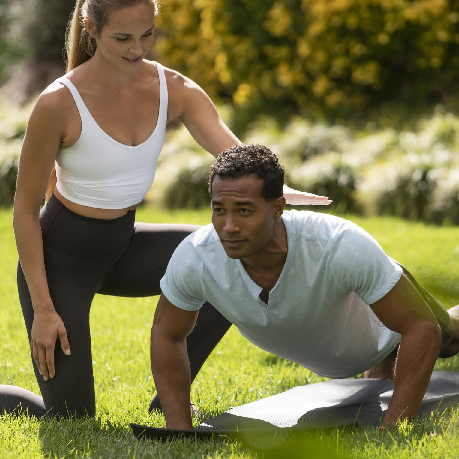 A yoga instructor helping a man get into a yoga position outside on very green grass.