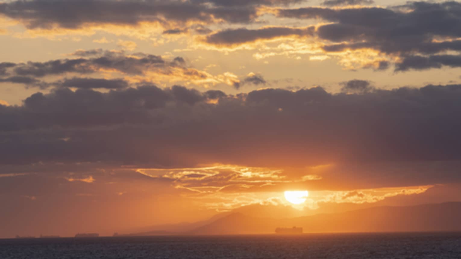 The sun setting behind the clouds and radiating an orange light ove the ocean below and sky above