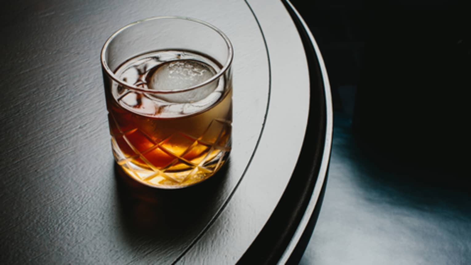 Liquor with large ice cube in rock glass on dark bar