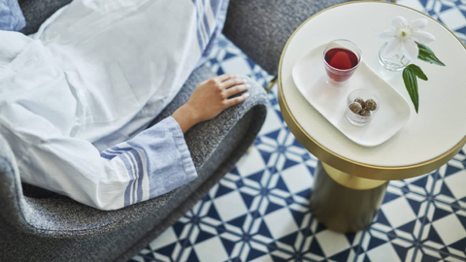 An aerial view of a woman relaxing in a spa lounge chair, a small bowl, a cup of dark red tea, and a white flower sit on a side table