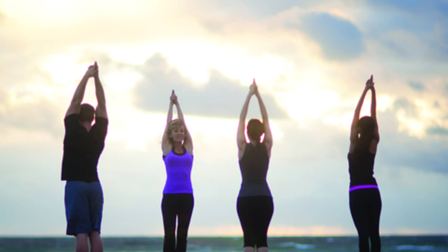 Four people stand in yoga poses with arms raised to the sky on beach at sunset