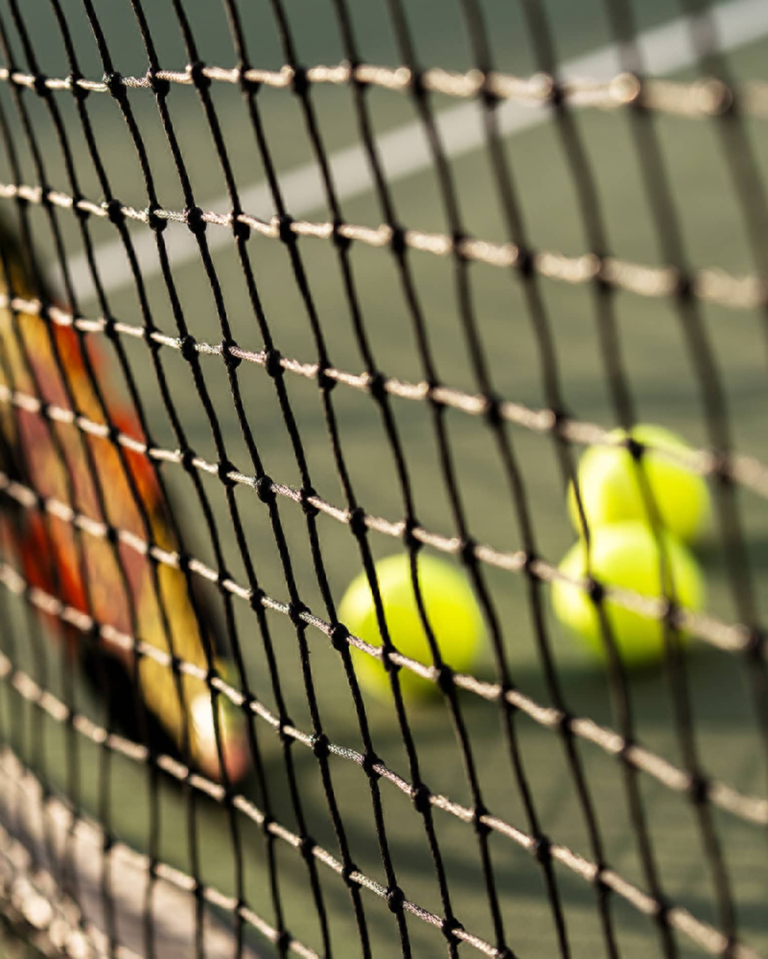 Close-up of tennis racket against net, three green tennis balls on ground