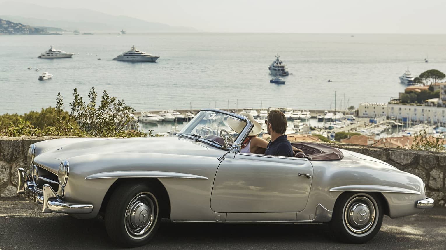 Couple sits in silver vintage convertible car parked on road by stone wall, looking over sea 