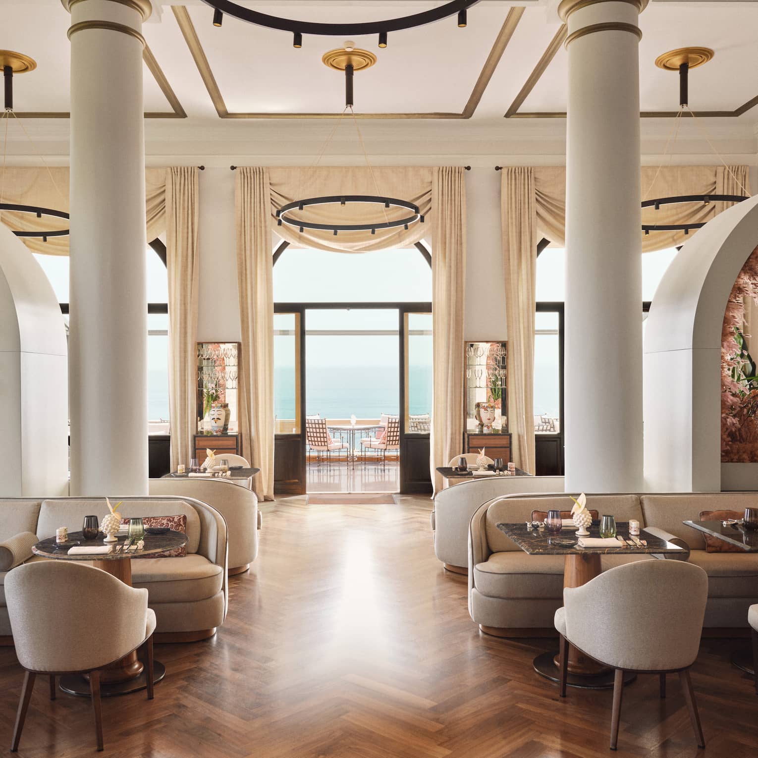 Restaurant dining room with two large columns in the centre, parquet wooden flooring, tall ceilings and a wall of windows that opens up to a terrace overlooking the sea