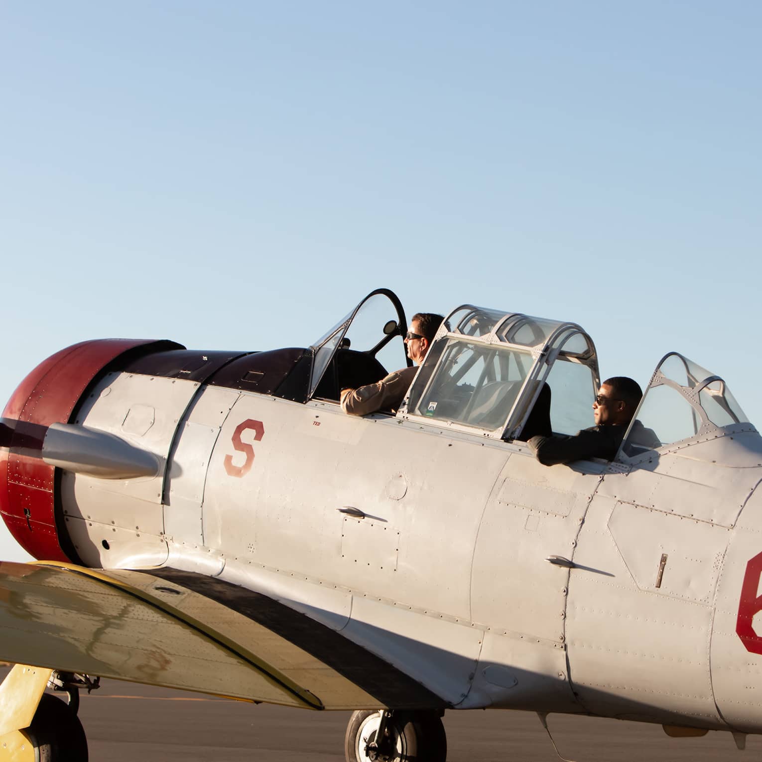 Two people sit in the cockpit of a vintage U.S. Navy aircraft on a runway, preparing for takeoff under a clear sky.