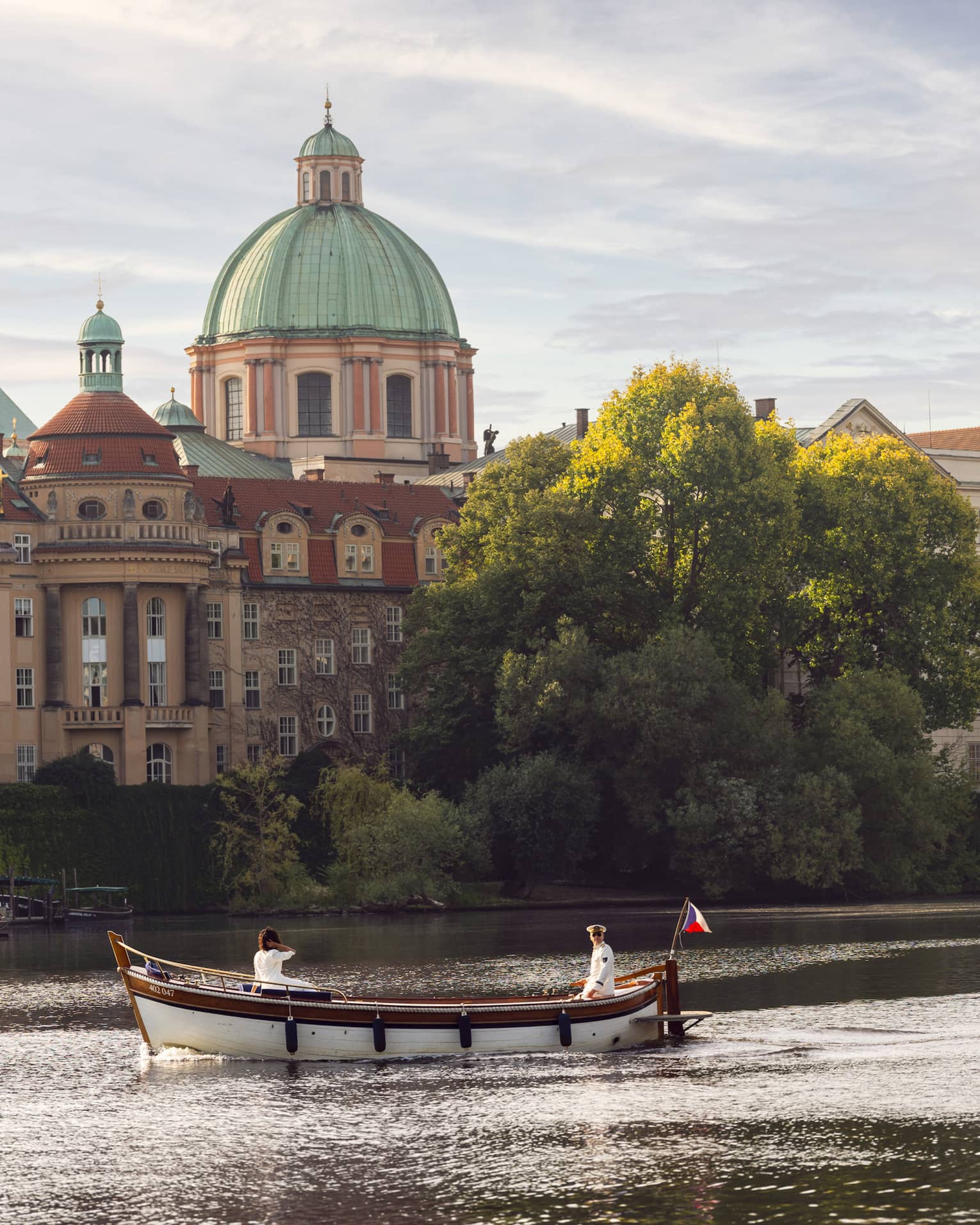 A boat with two people sailing on a river, with historic buildings, including a large dome and a tower, in the background.
