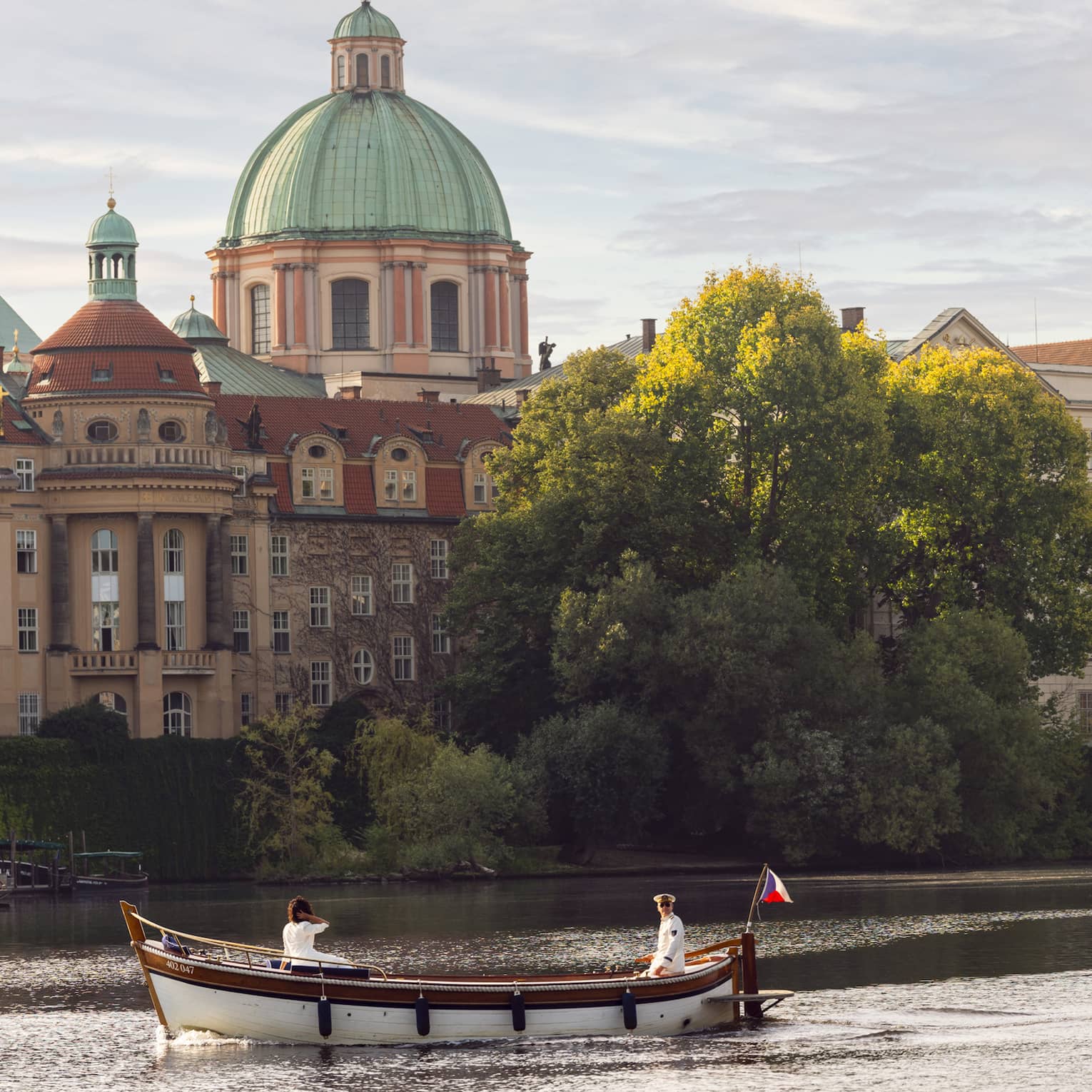 A boat with two people sailing on a river, with historic buildings, including a large dome and a tower, in the background.