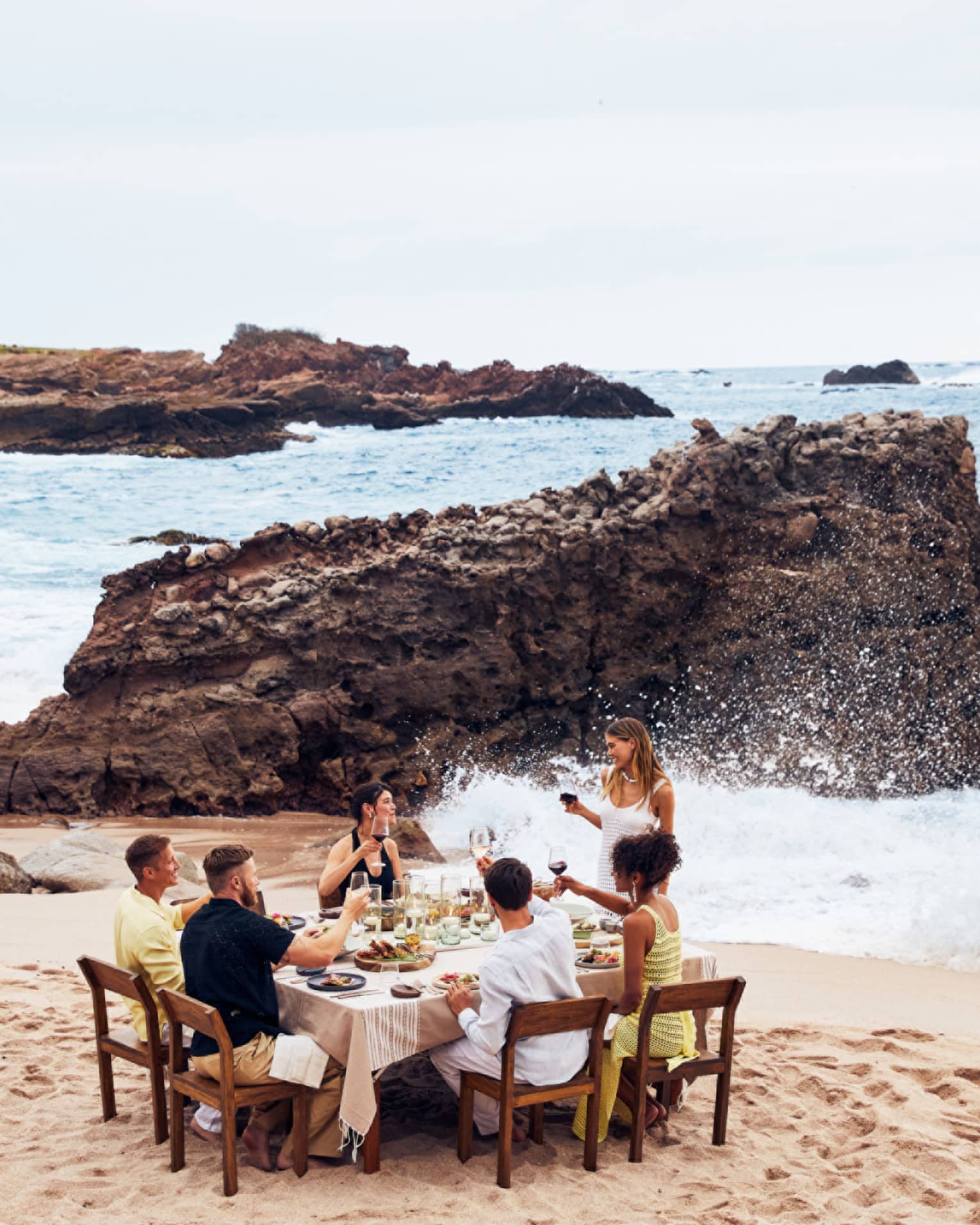 A group eating at a table on a beach with waves crashing nearby.