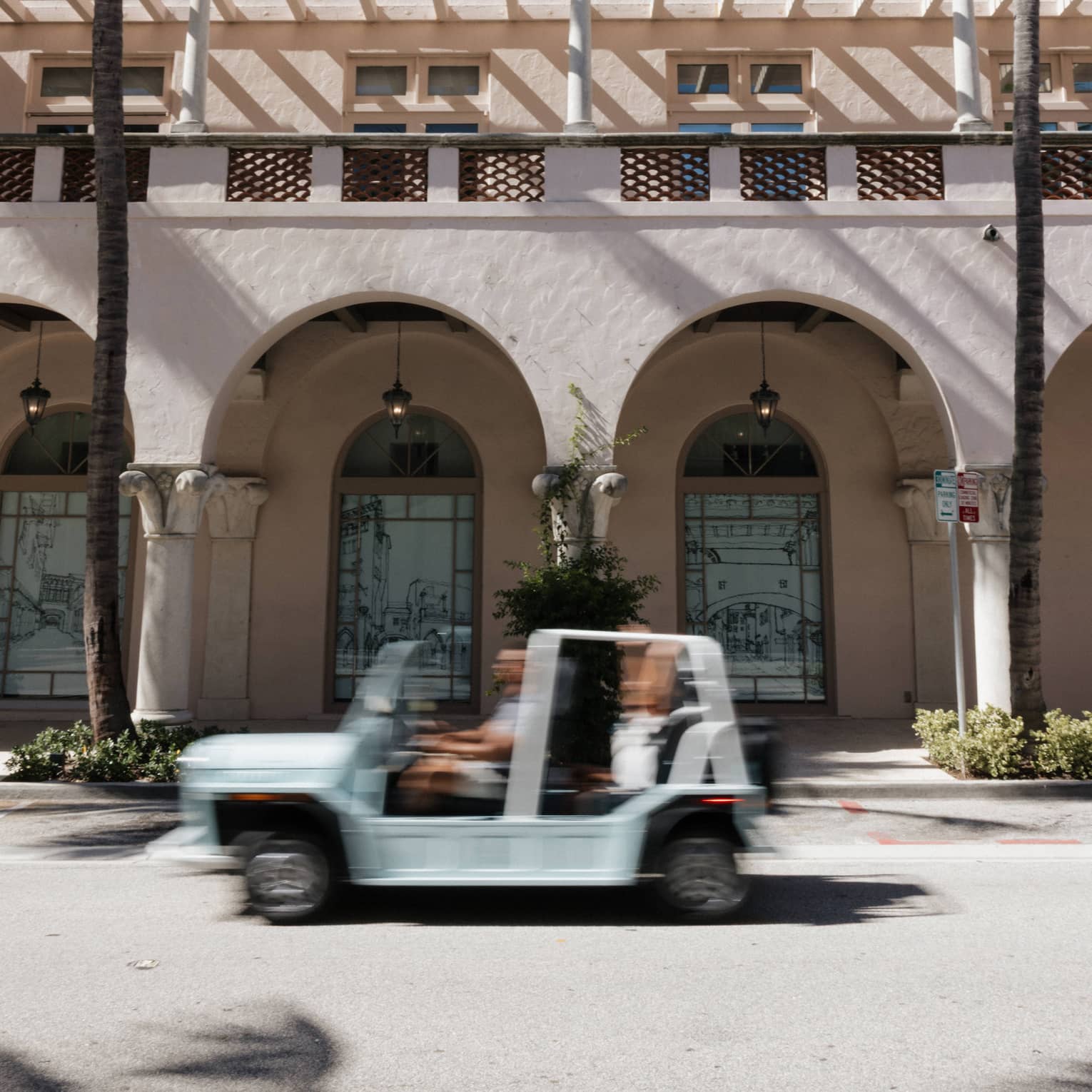 White open-top vehicle drives by a Spanish-style stucco building with palm trees in front