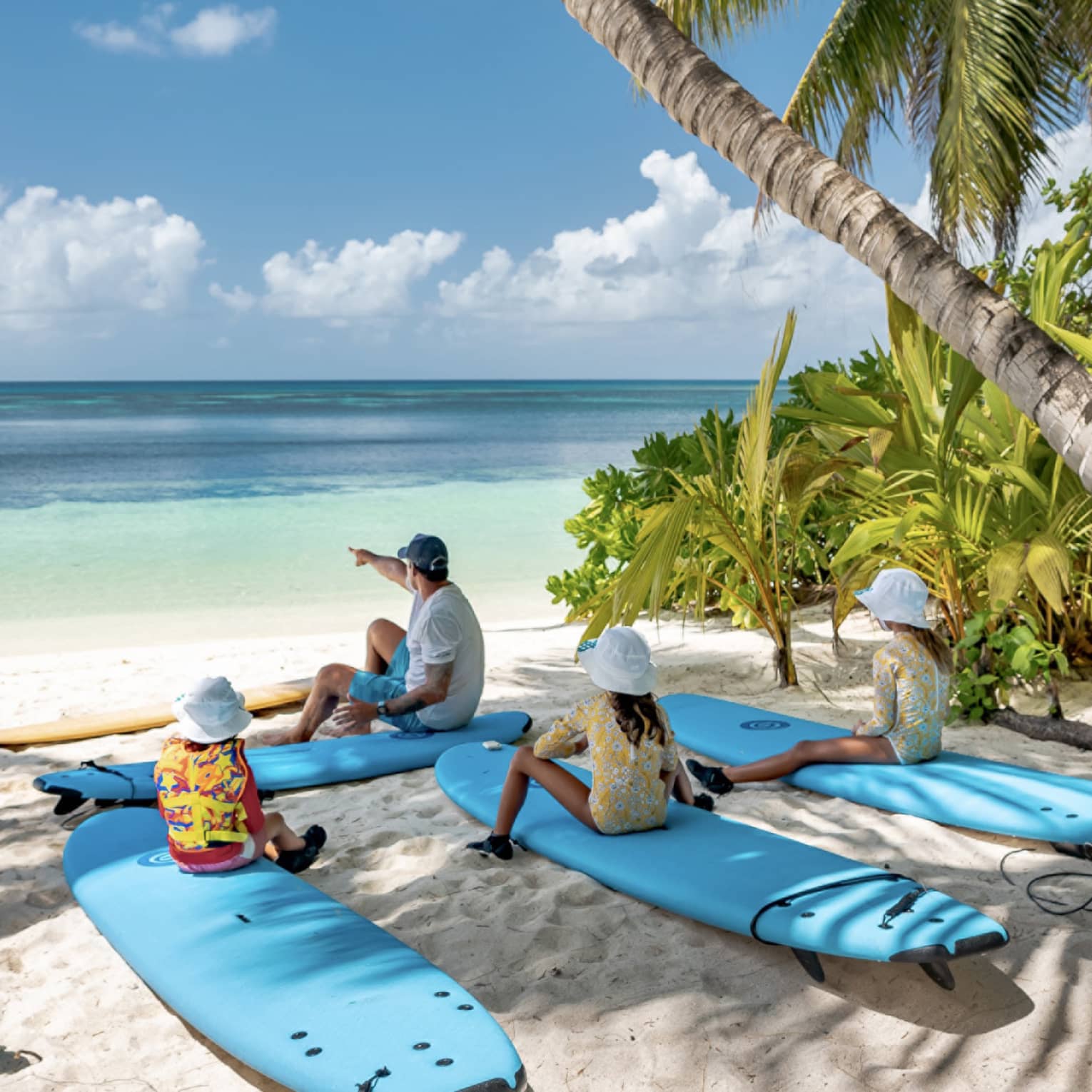 Children sit on surfboards under the shade of palm trees at the beach. One of their two instructors point at the clear water.