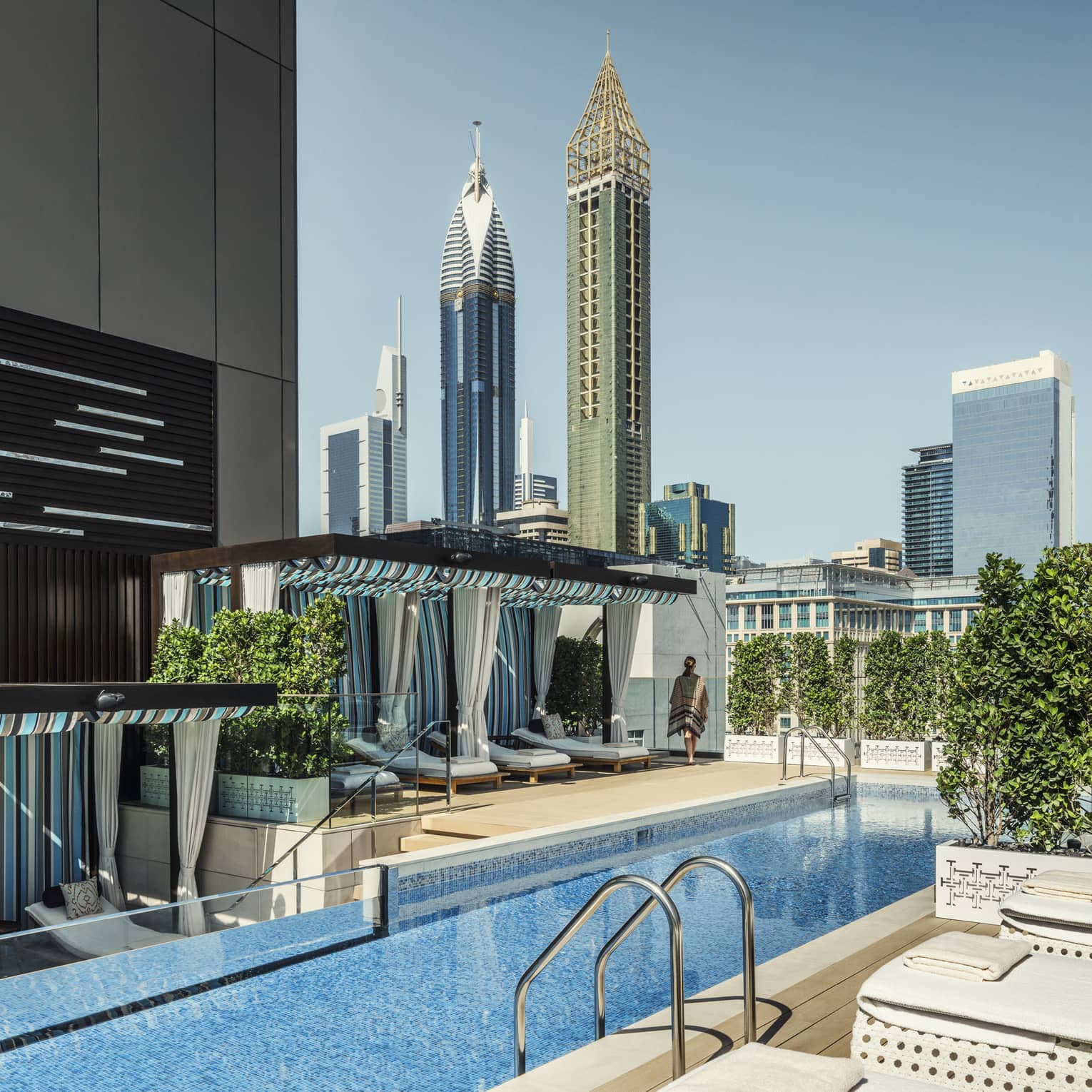 White lounge chairs, ladder into long blue swimming pool by cabanas on rooftop patio on sunny day