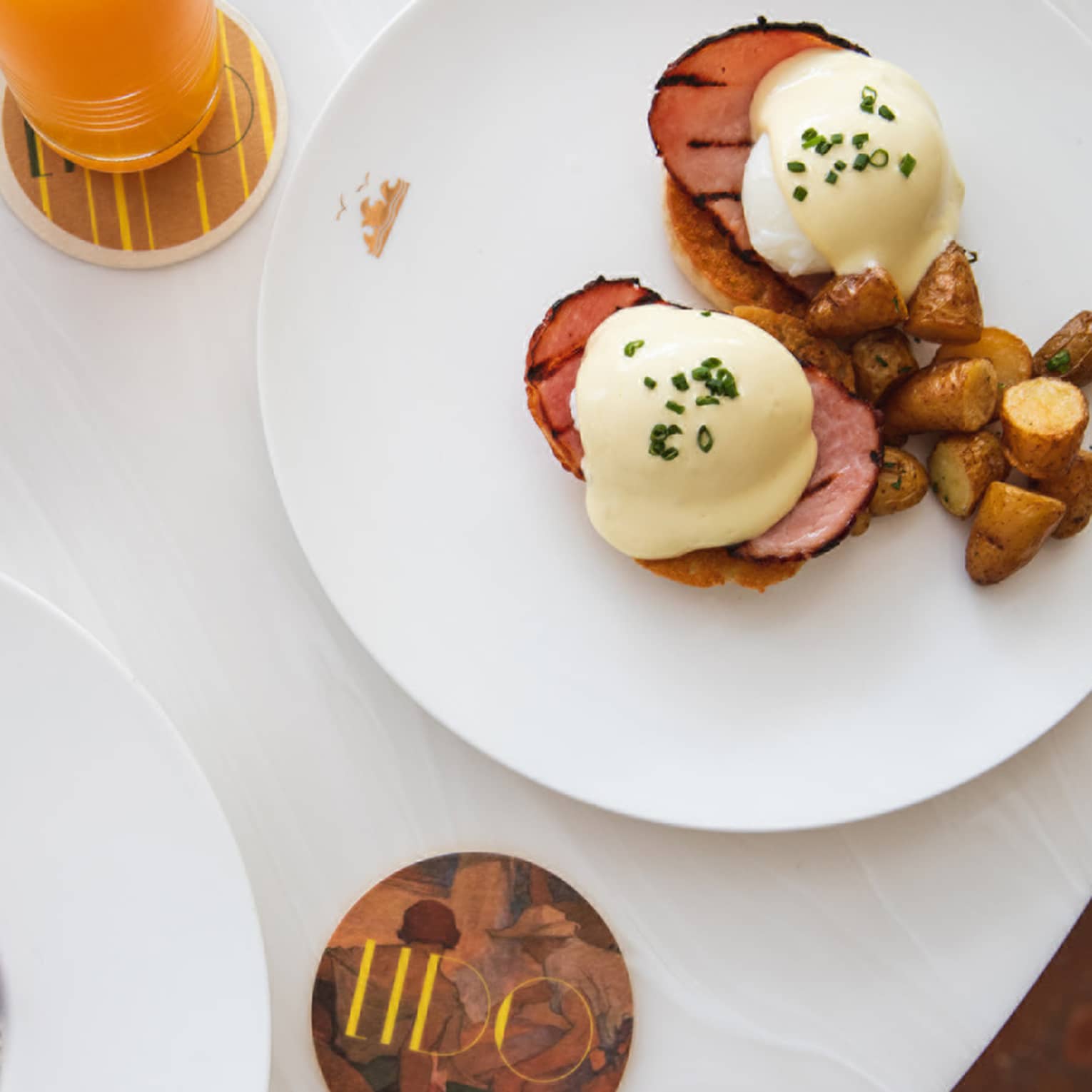 Two plates, one with eggs Benedict and a side of roasted potatoes and one with chia seed pudding topped with berries, sit on a white marble table next to an orange flower in a vase