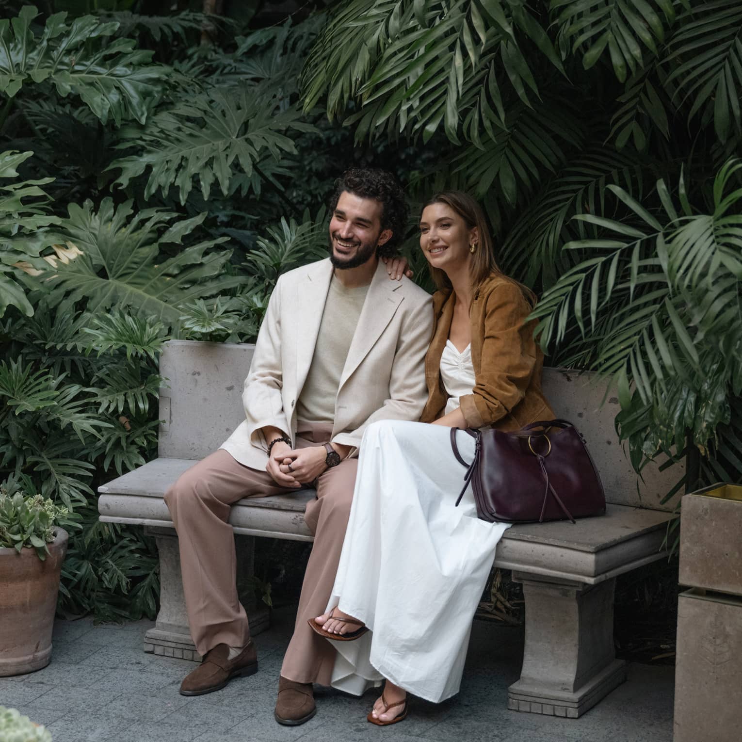 Two people sit on a bench outdoors, surrounded by greenery and plantings.