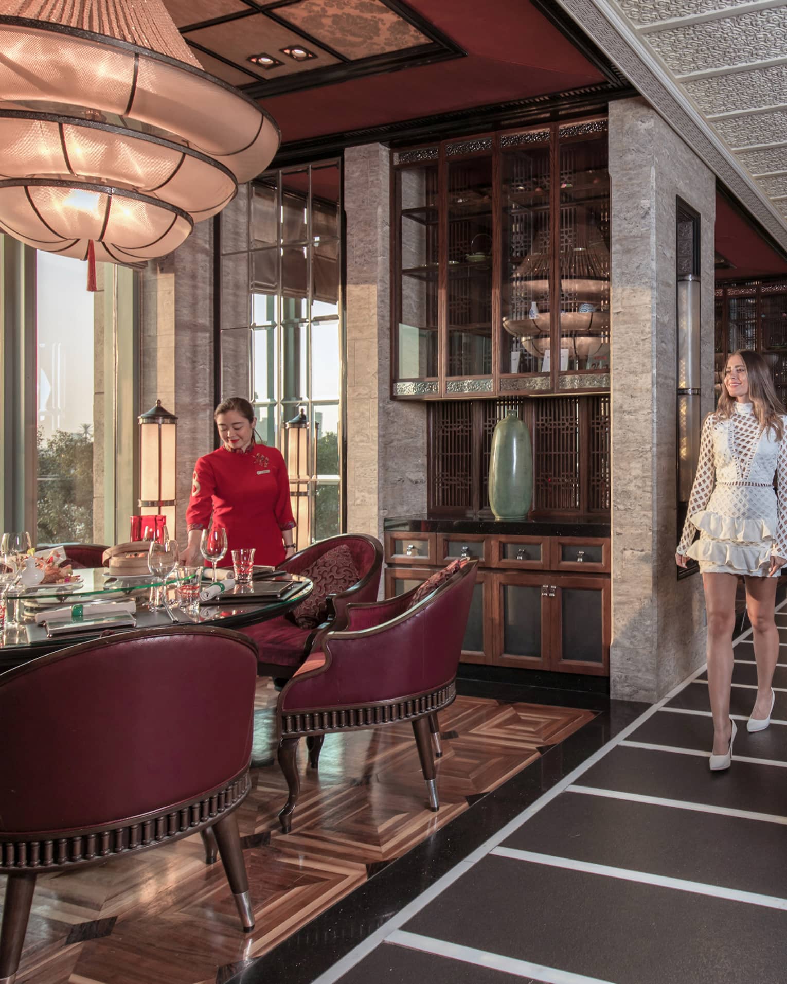 A guest is shown to her table, surrounded by burgundy chairs, in a luxurious restaurant with opulent decorations. 