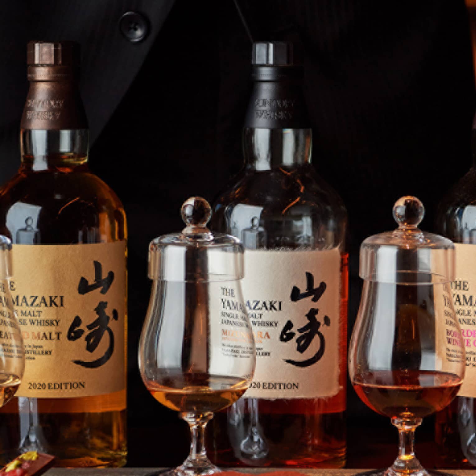 Hands frame a row of three bottles of single malt Japanese whisky behind three curved whisky tumblers with tasting caps.