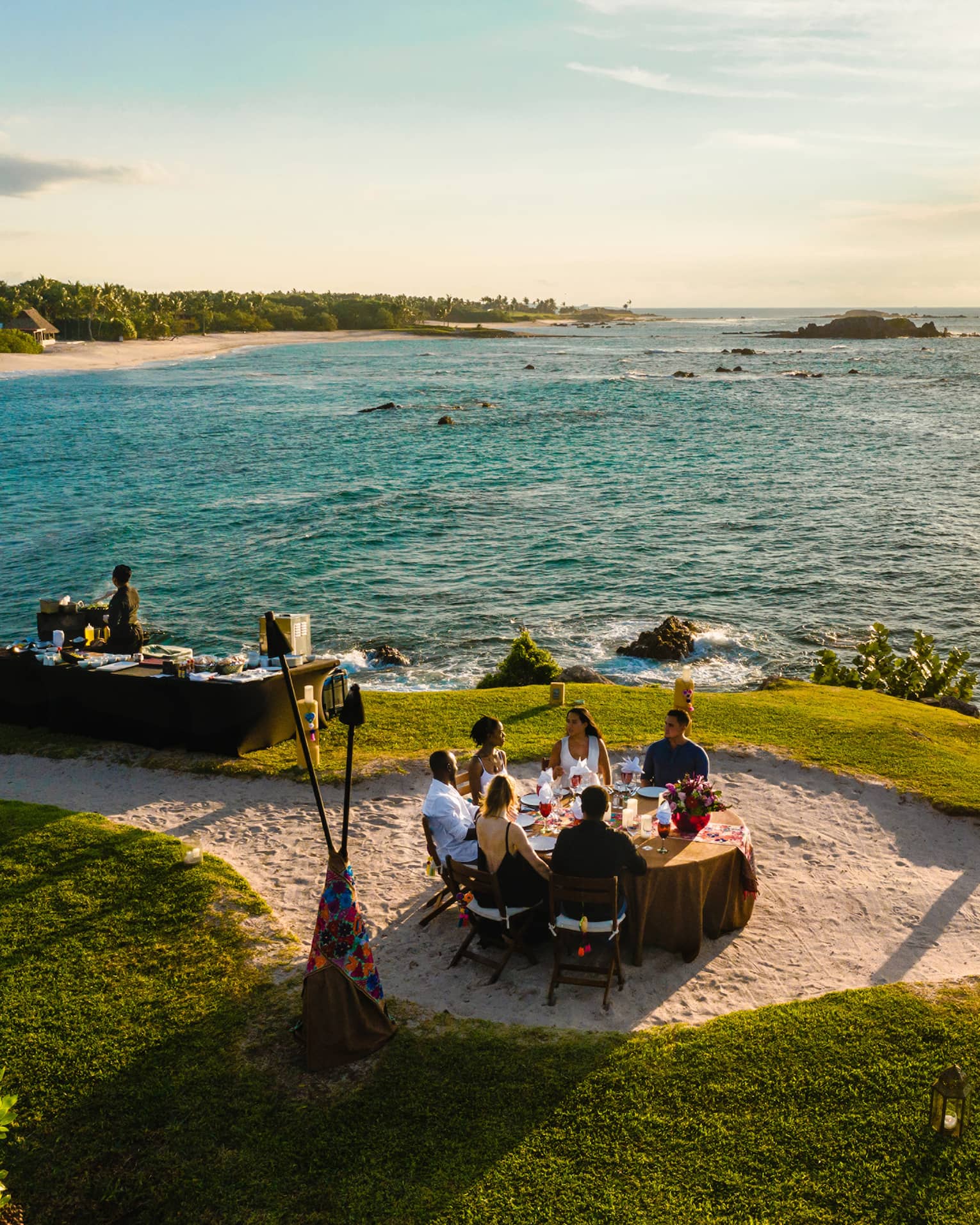 Aerial view of Dinner on the Rock dining table on lawn, sandy path near ocean at sunset