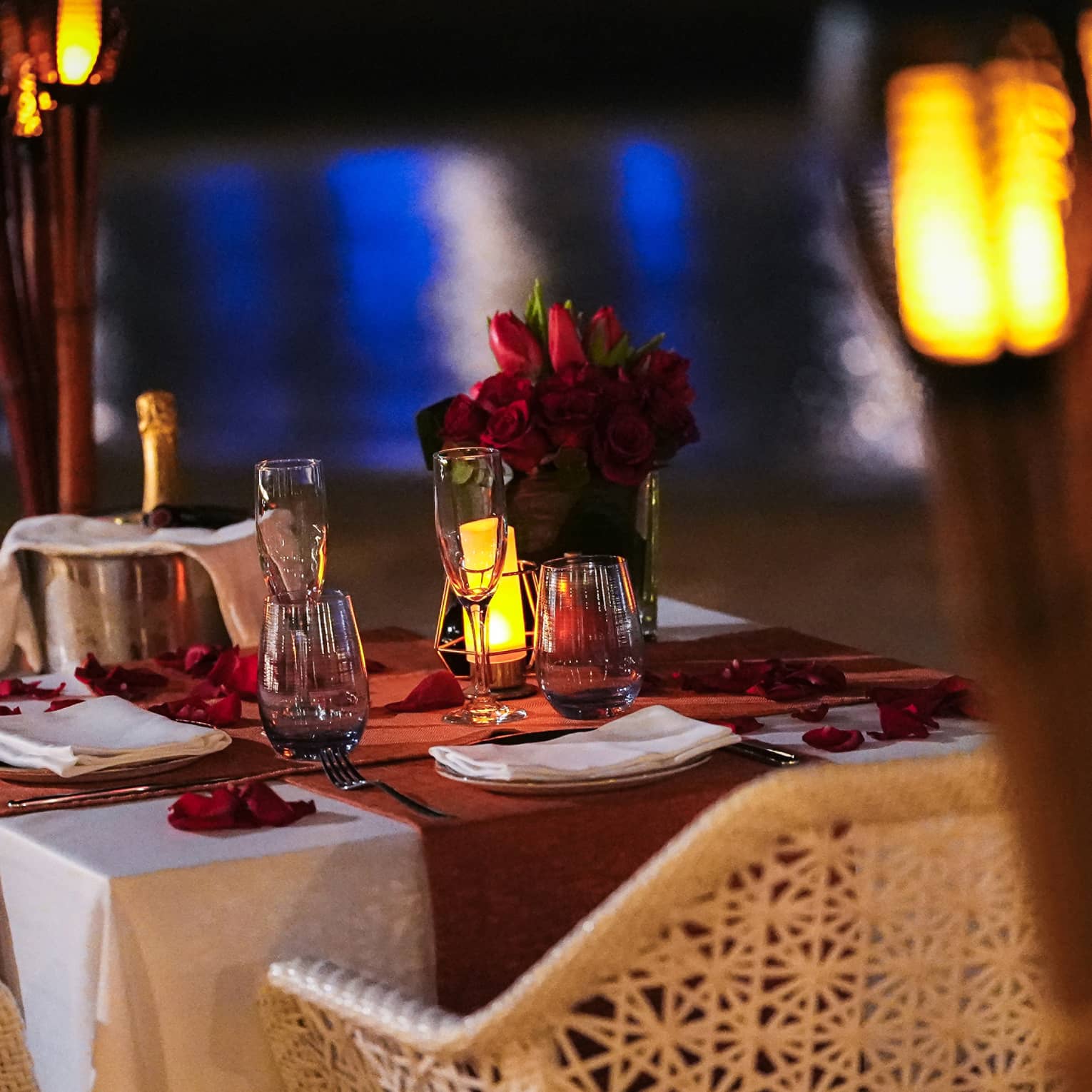 Table for two set with chilling champagne, flutes and a rose bouquet, aside torches and the gleaming reflection on the water.