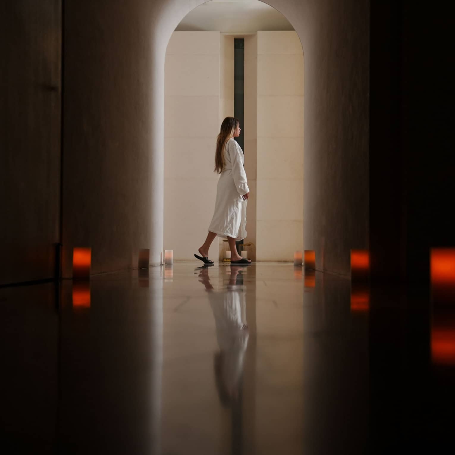 Person with long dark hair wearing a white spa robe walks through a dark hallway lit by eight dim red lanterns