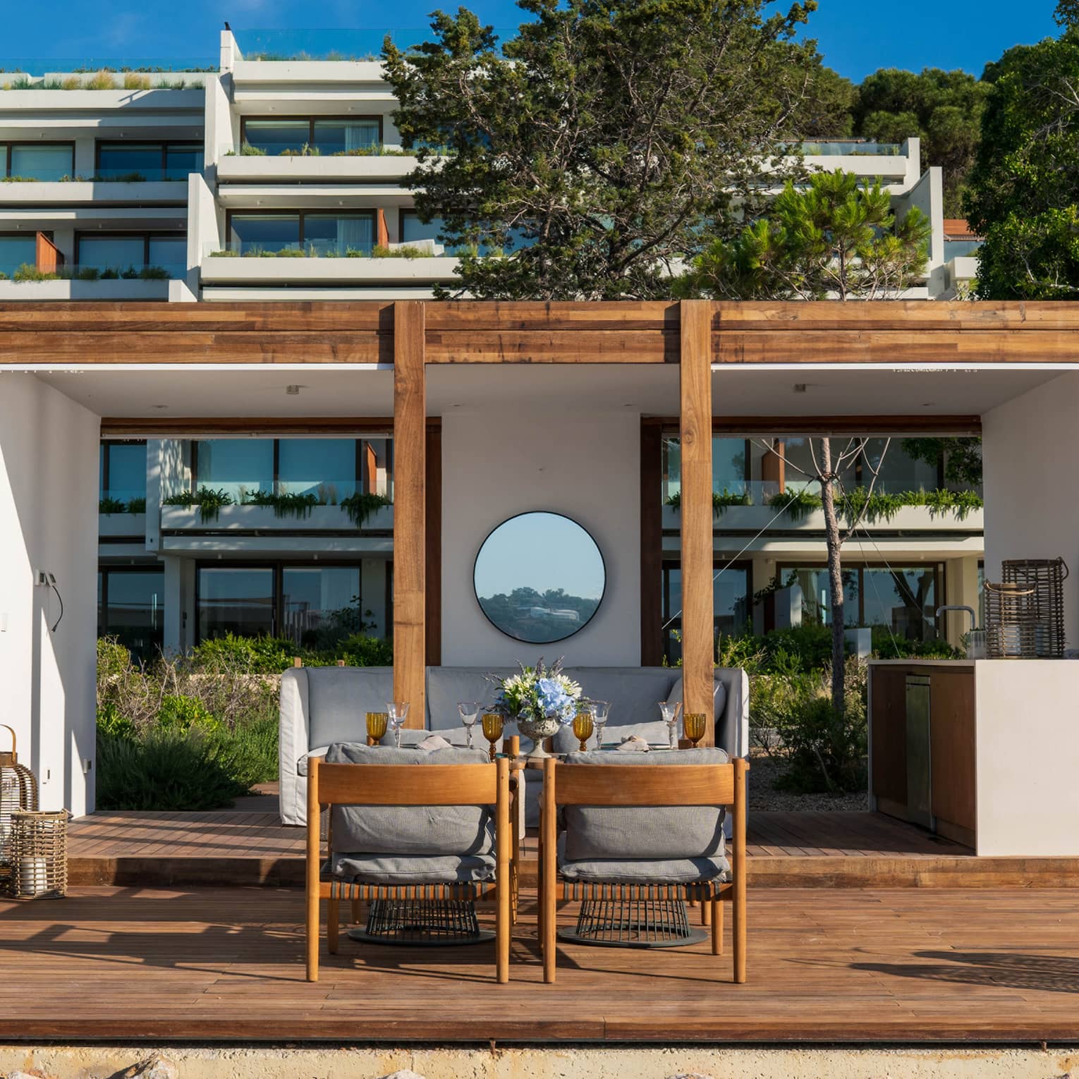 A pristine white cabana with wood accents frames a patio dining area in front of hotel balconies laden with greenery. 