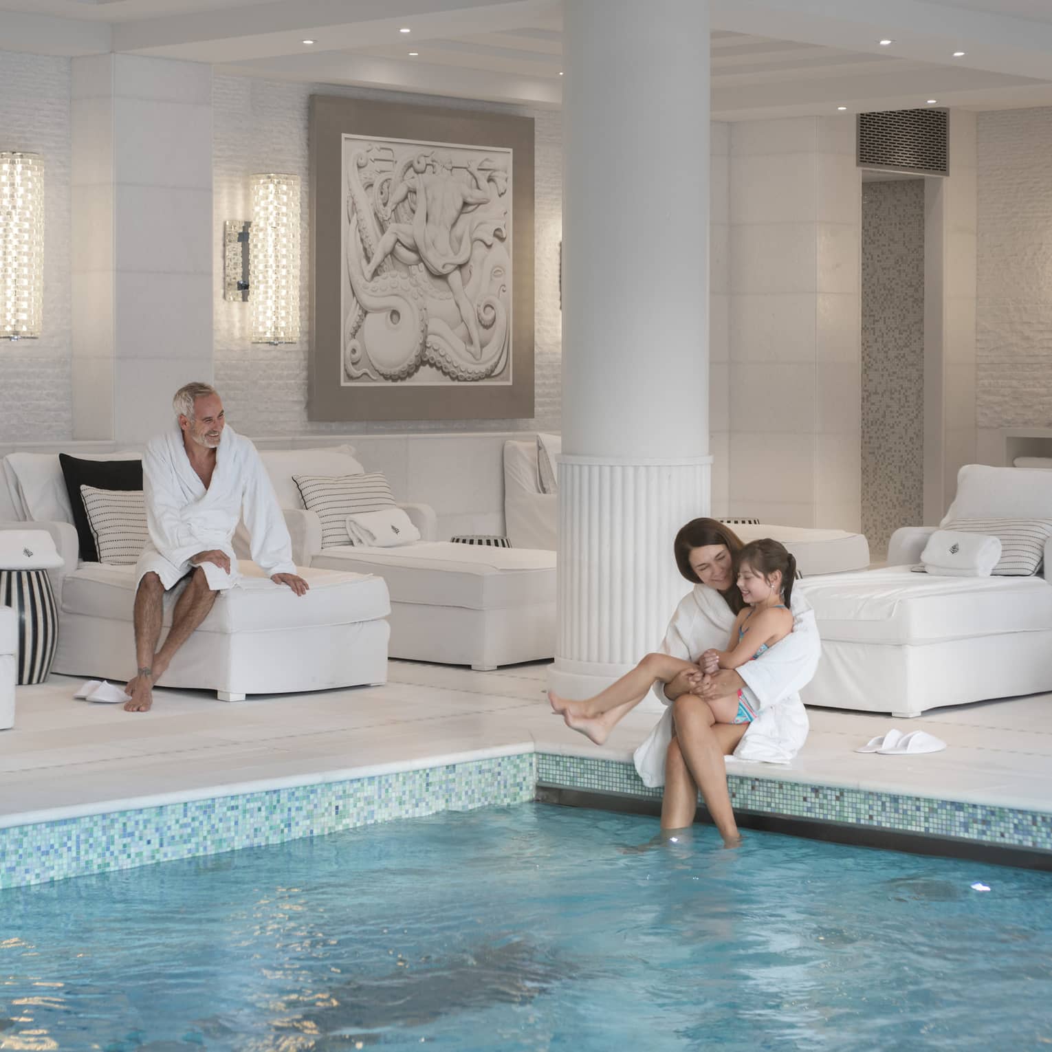 A child sits on her mother’s lap at the edge of an indoor pool as another guest smiles at them from a plush lounge chair.